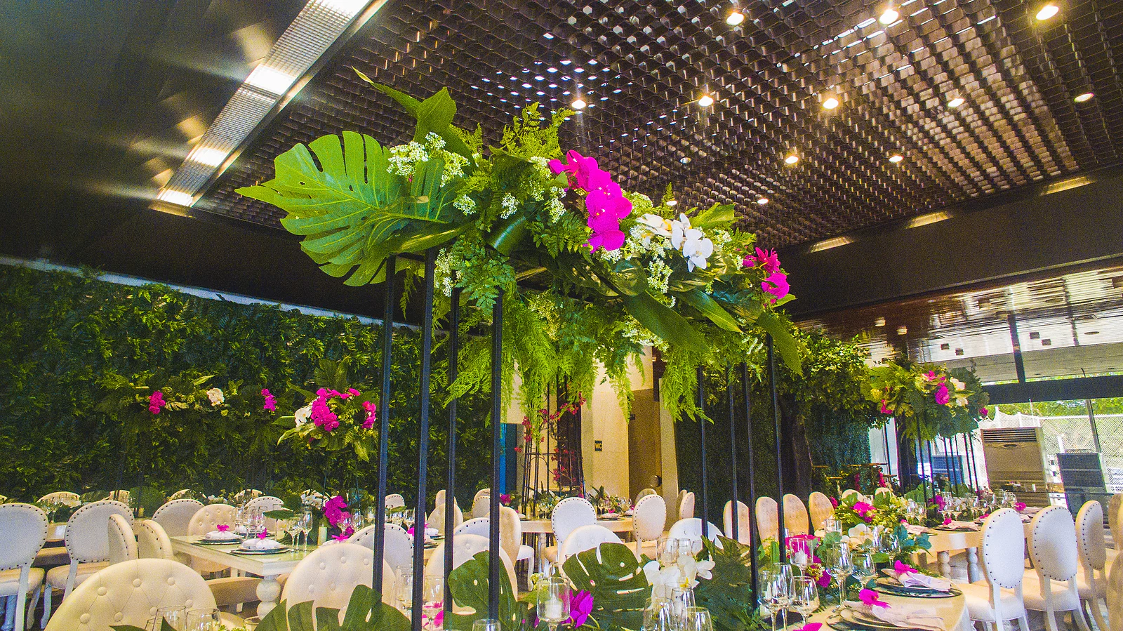 Centro de mesa alto, sobre pés pretos, com folhas de monstera, fetos e orquídeas fúcsia e brancas, junto às mesas postas