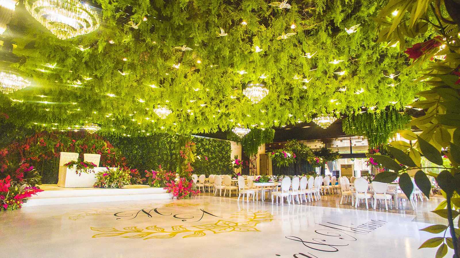 Salão com o tecto coberto de folhagem suspensa e luzes, pista branca com monograma dourado e preto e a mesa dos noivos ao fundo