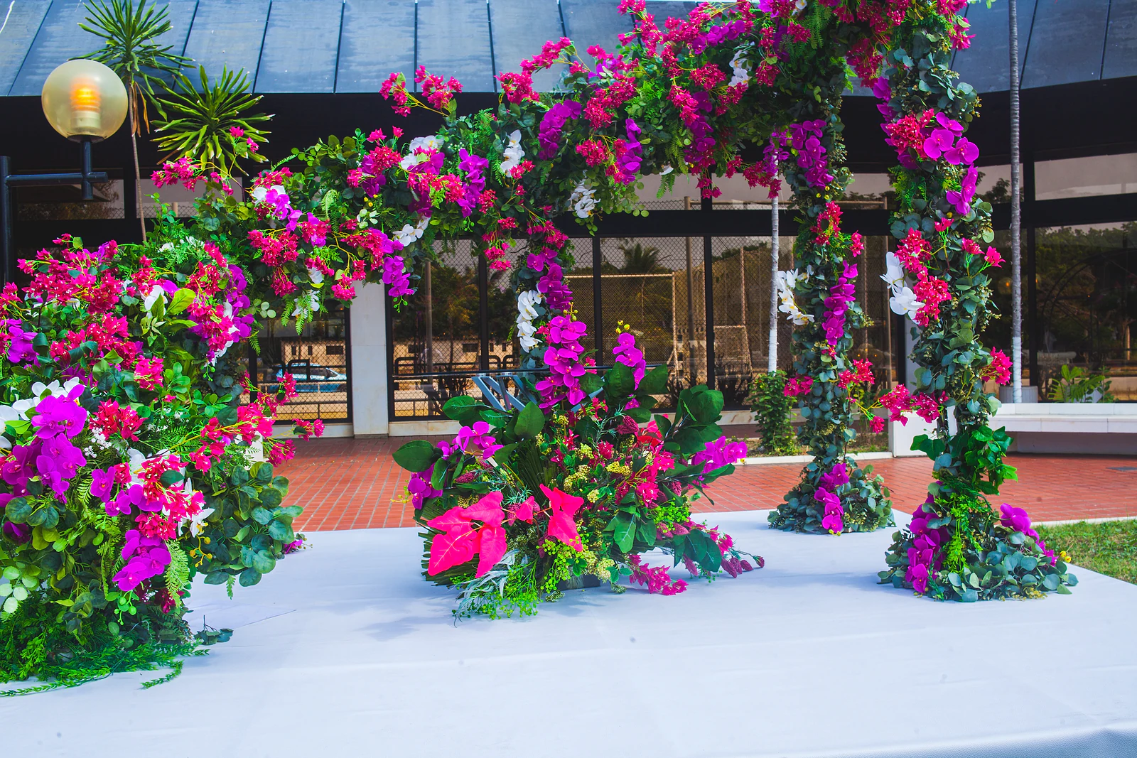 Pérgola coberta de flores fúcsia e roxas sobre um estrado branco, com um candeeiro de globos ao lado