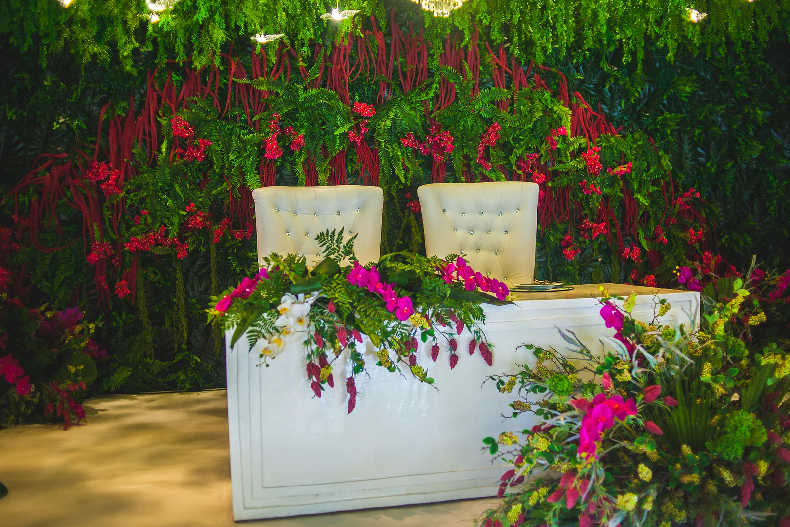 Pormenor da mesa dos noivos: duas cadeiras brancas capitonadas, um ramo de orquídeas fúcsia e folhagem sobre a frente da mesa e um arranjo de folhagem e flores à direita