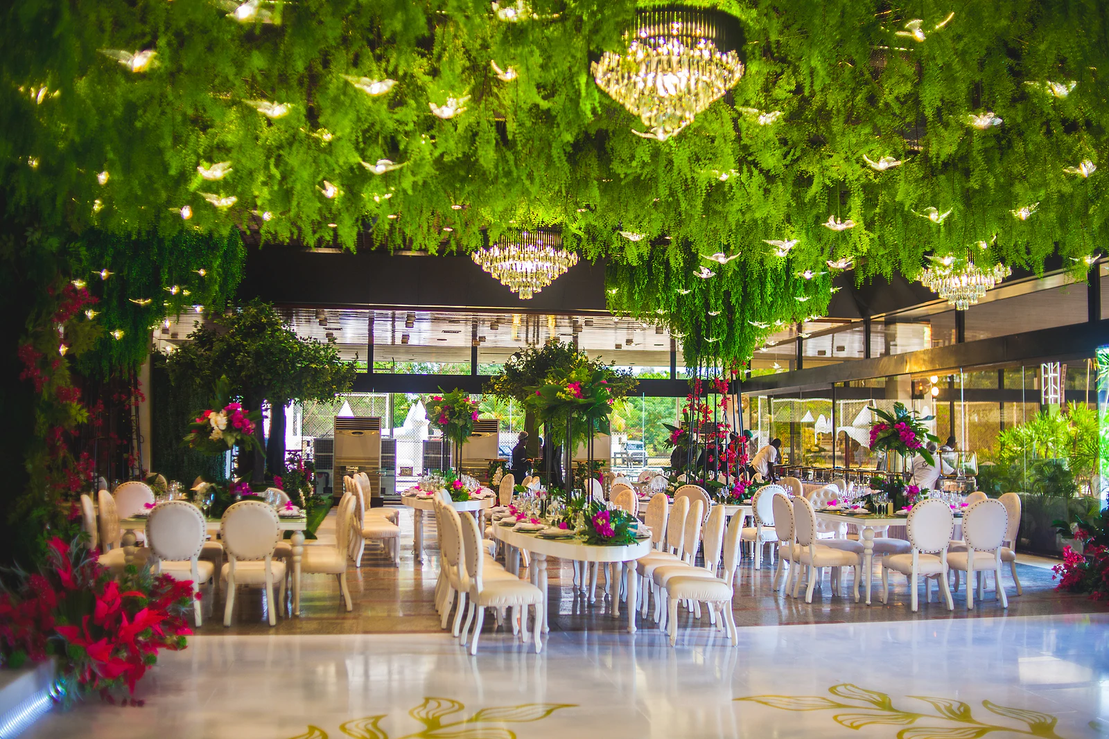 Salão com mesas compridas postas, cadeiras brancas e centros altos de flores, sob um tecto de folhagem e lustres de cristal