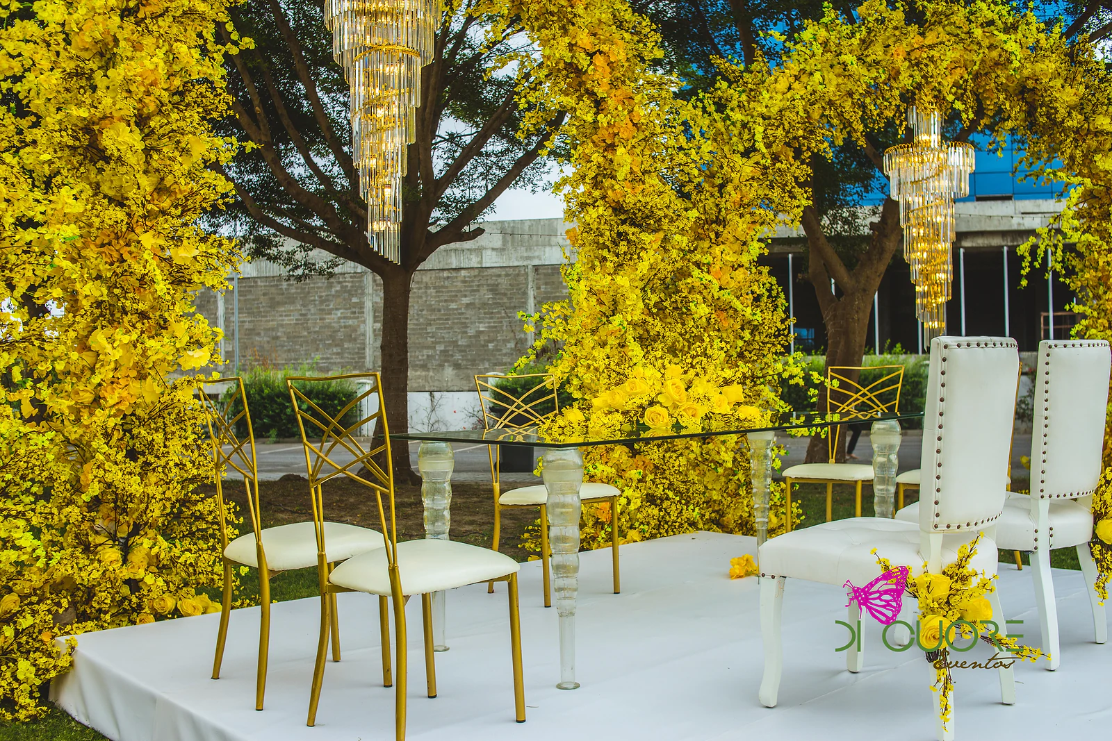 Estrado branco com mesa de vidro, cadeiras douradas e cadeiras brancas capitonadas, sob um arco de flores amarelas com pendentes de cristal