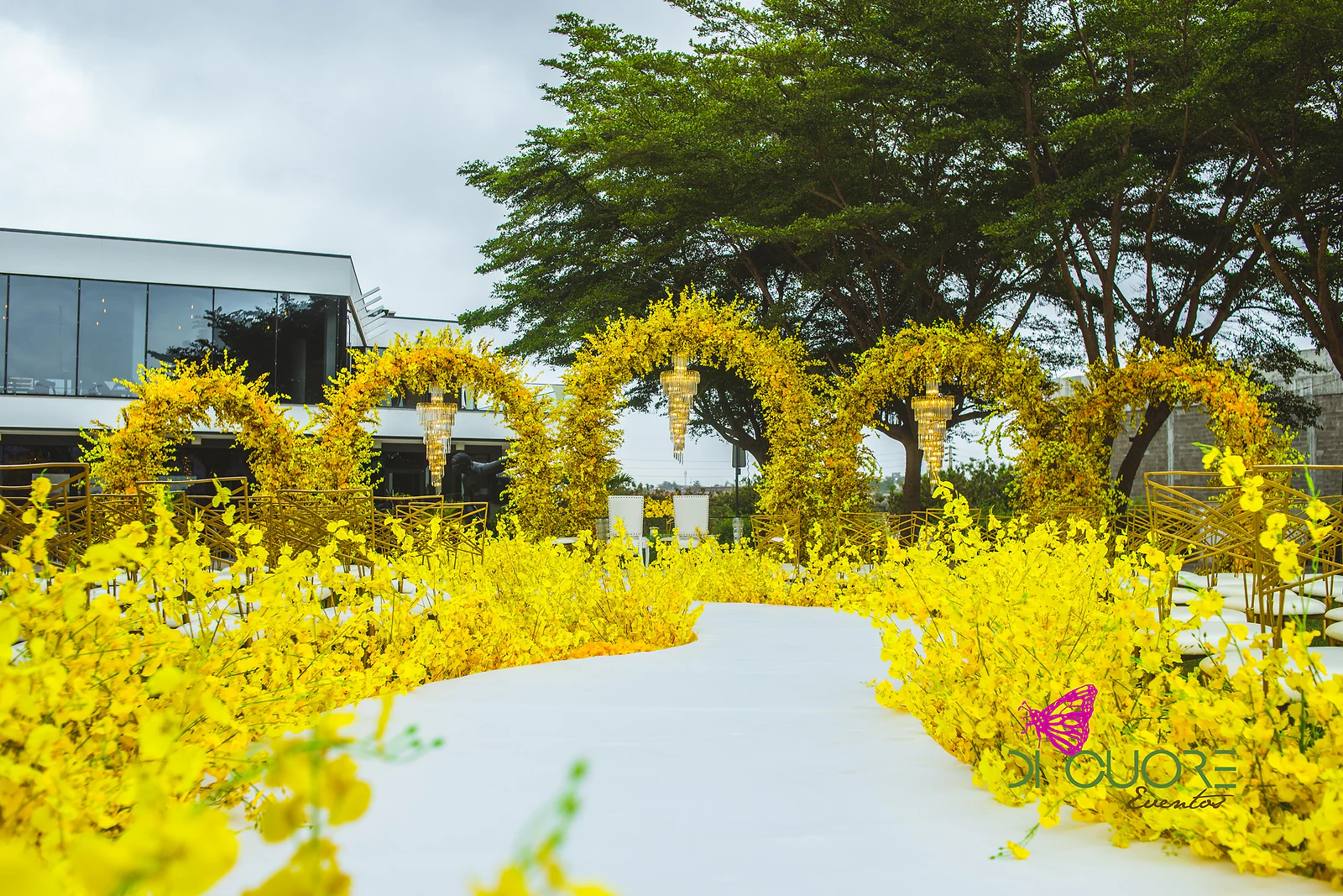 Passadeira branca em curva entre maciços de flores amarelas, com uma fila de arcos de flores amarelas e lustres suspensos, diante de um edifício moderno e de uma grande árvore