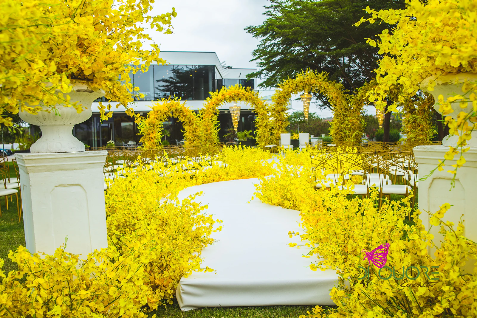 Passadeira branca entre maciços de flores amarelas e dois pedestais brancos com vasos de flores amarelas, com arcos floridos ao fundo