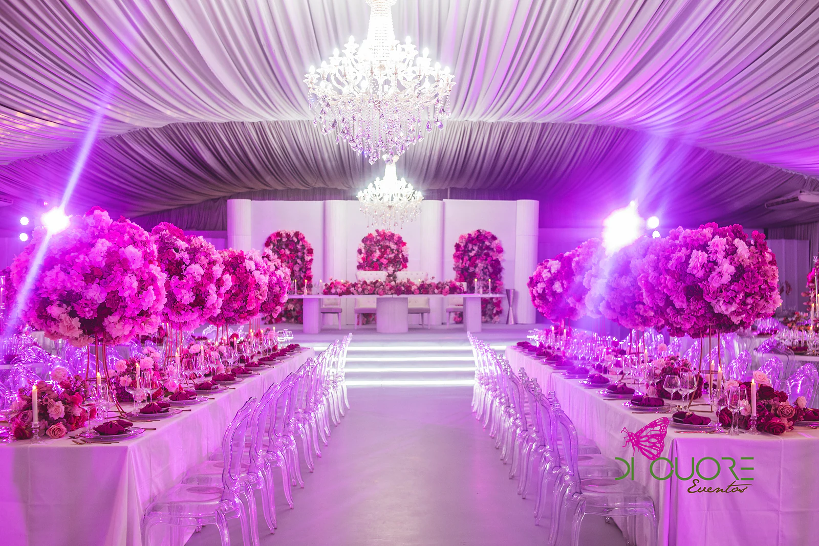 Corredor central do salão entre duas mesas corridas com bolas de flores fúcsia, cadeiras transparentes e lustres de cristal, com a mesa principal ao fundo