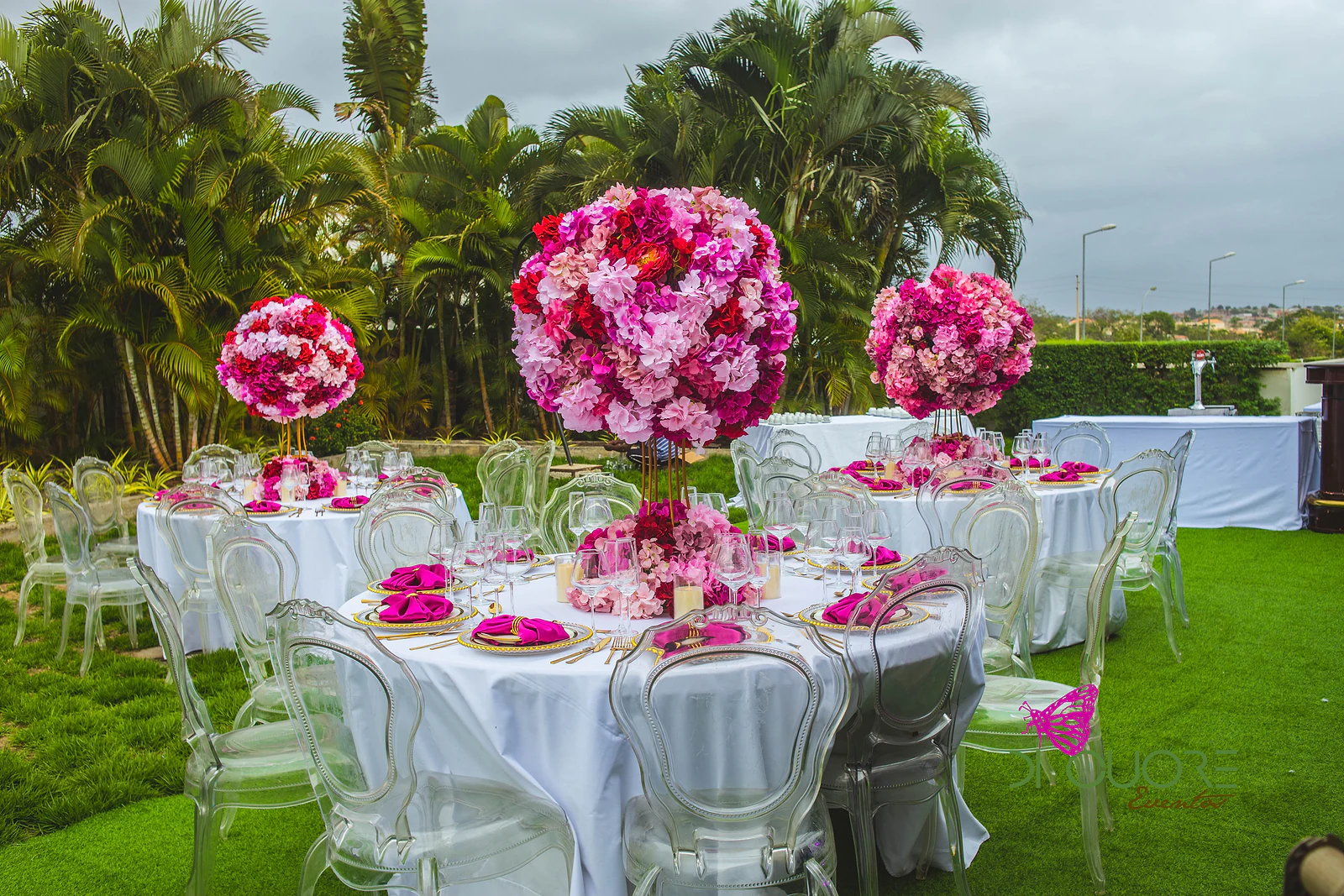 Mesas redondas de toalha branca e cadeiras transparentes no relvado, com bolas de flores cor-de-rosa e vermelhas como centros de mesa, entre palmeiras