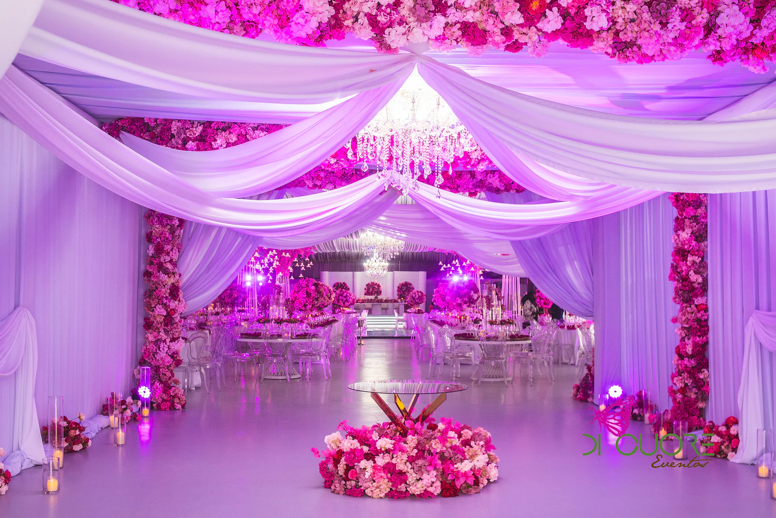 Entrada do salão com cortinados em festões brancos, pilares de flores cor-de-rosa e um arranjo redondo de flores no chão, com as mesas em luz violeta ao fundo