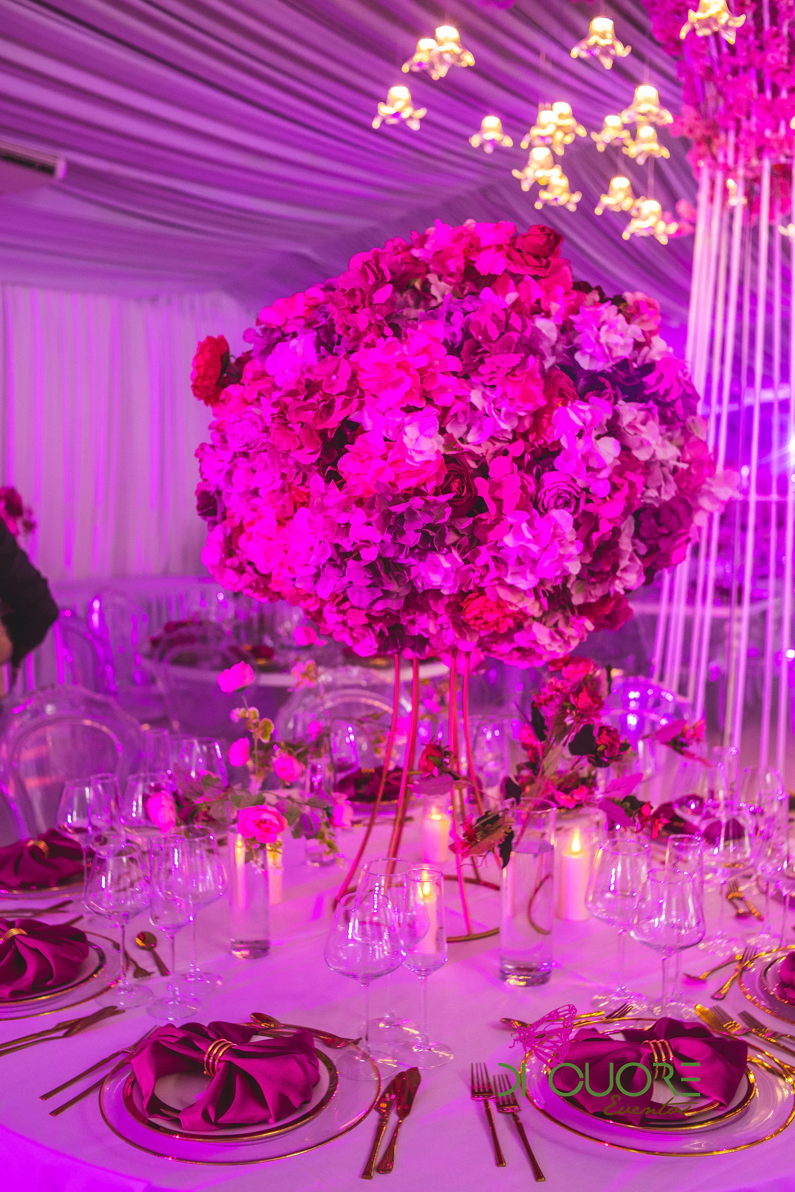 Grande bola de flores cor-de-rosa e fúcsia sobre pés dourados, rodeada de velas, copos de cristal e guardanapos fúcsia em laço