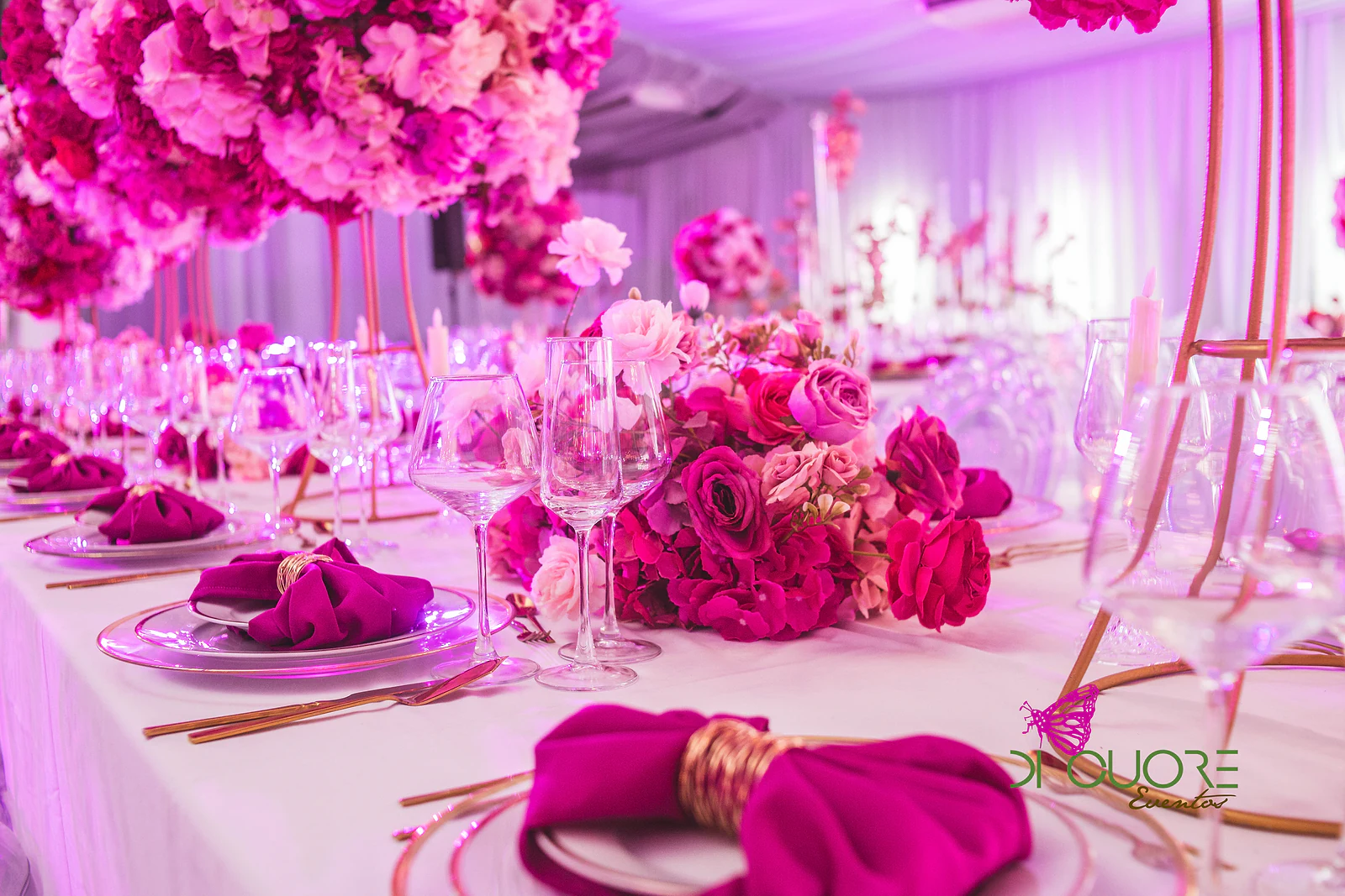 Mesa posta com guardanapos fúcsia em laço, talheres dourados e um ramo de rosas cor-de-rosa e vermelhas junto a copos de cristal