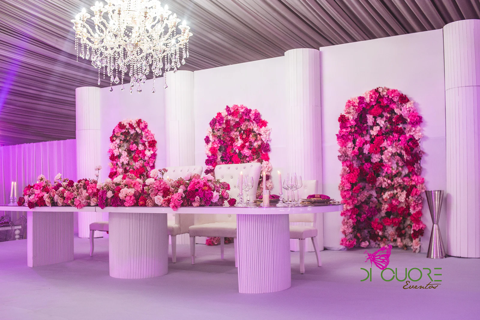 Mesa principal sobre pedestais brancos redondos, coberta de flores cor-de-rosa e vermelhas, diante de painéis brancos com arcos de flores e um lustre de cristal