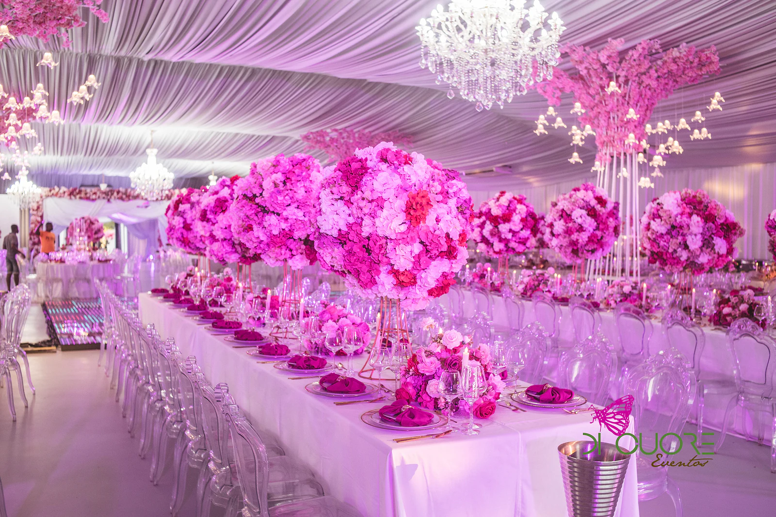 Mesa corrida com toalha branca, cadeiras transparentes e grandes bolas de flores cor-de-rosa, sob um lustre de cristal e uma árvore de flores suspensa