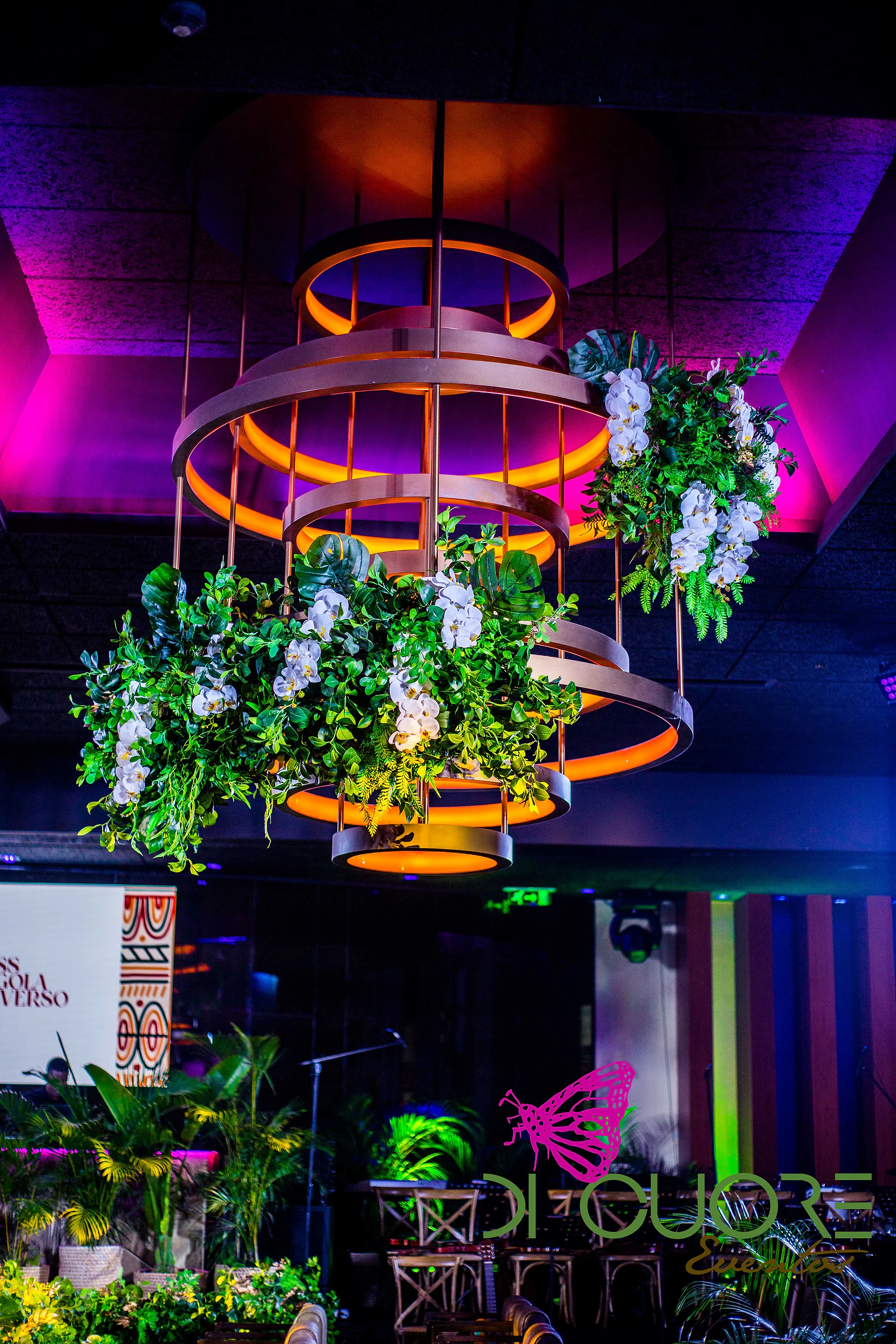 Lustre de aros dourados com cachos de folhagem e orquídeas brancas suspensos, sob luz magenta e laranja, sobre o salão