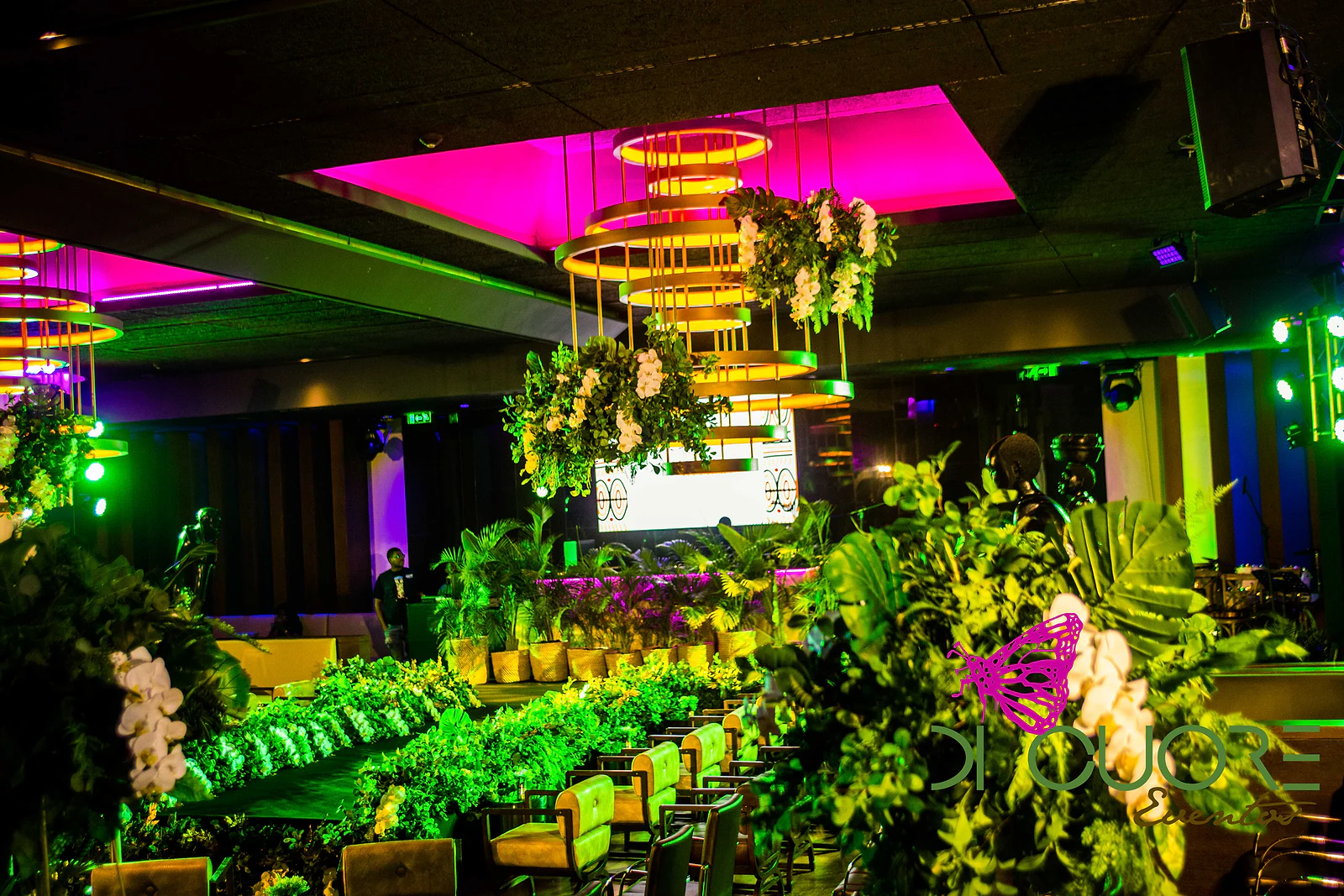Salão com folhagem e mesas iluminadas a verde e amarelo, lustres dourados com flores e tecto iluminado a magenta