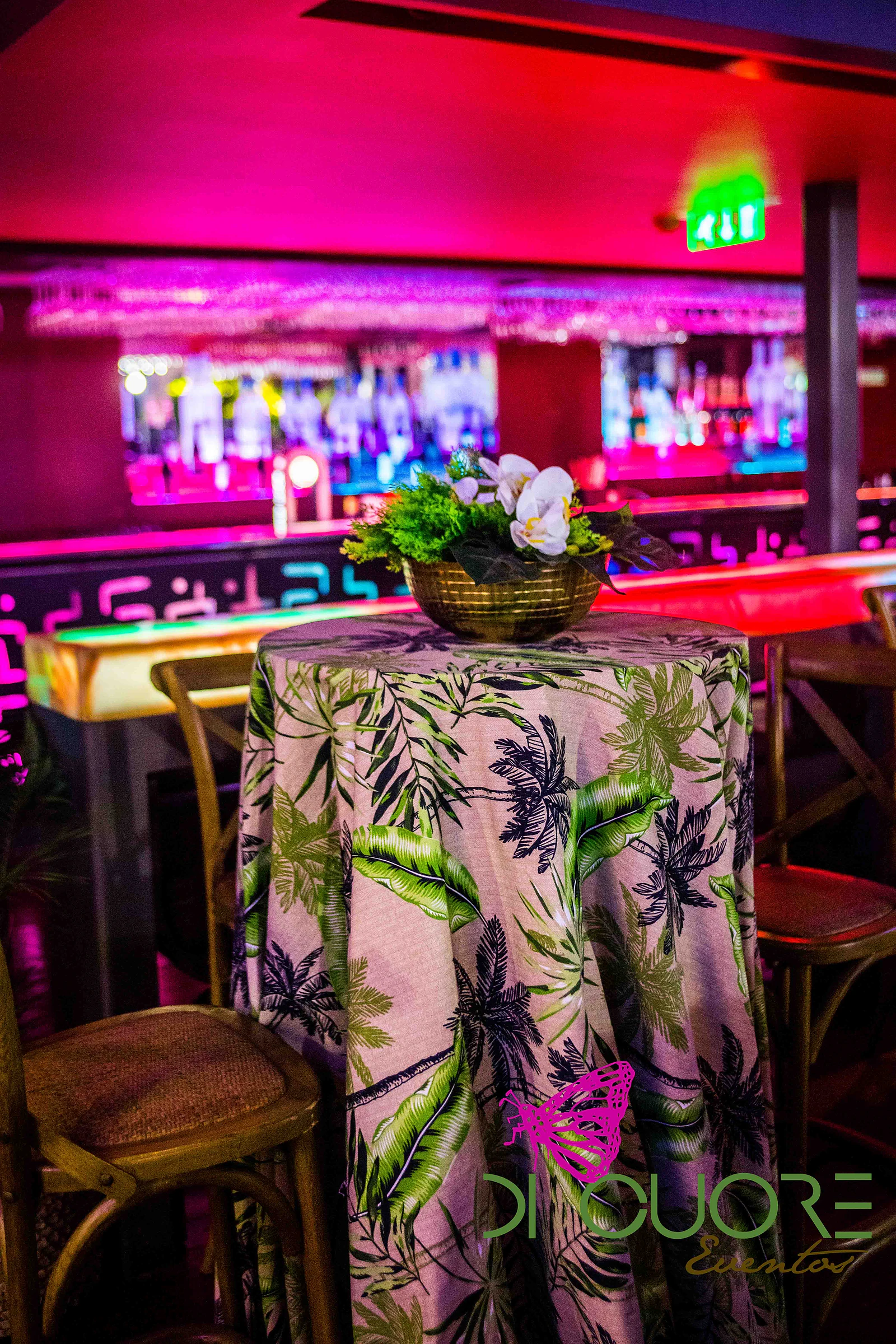 Mesa alta com toalha estampada de folhas de palmeira e uma taça dourada com orquídeas brancas, diante do bar iluminado a magenta