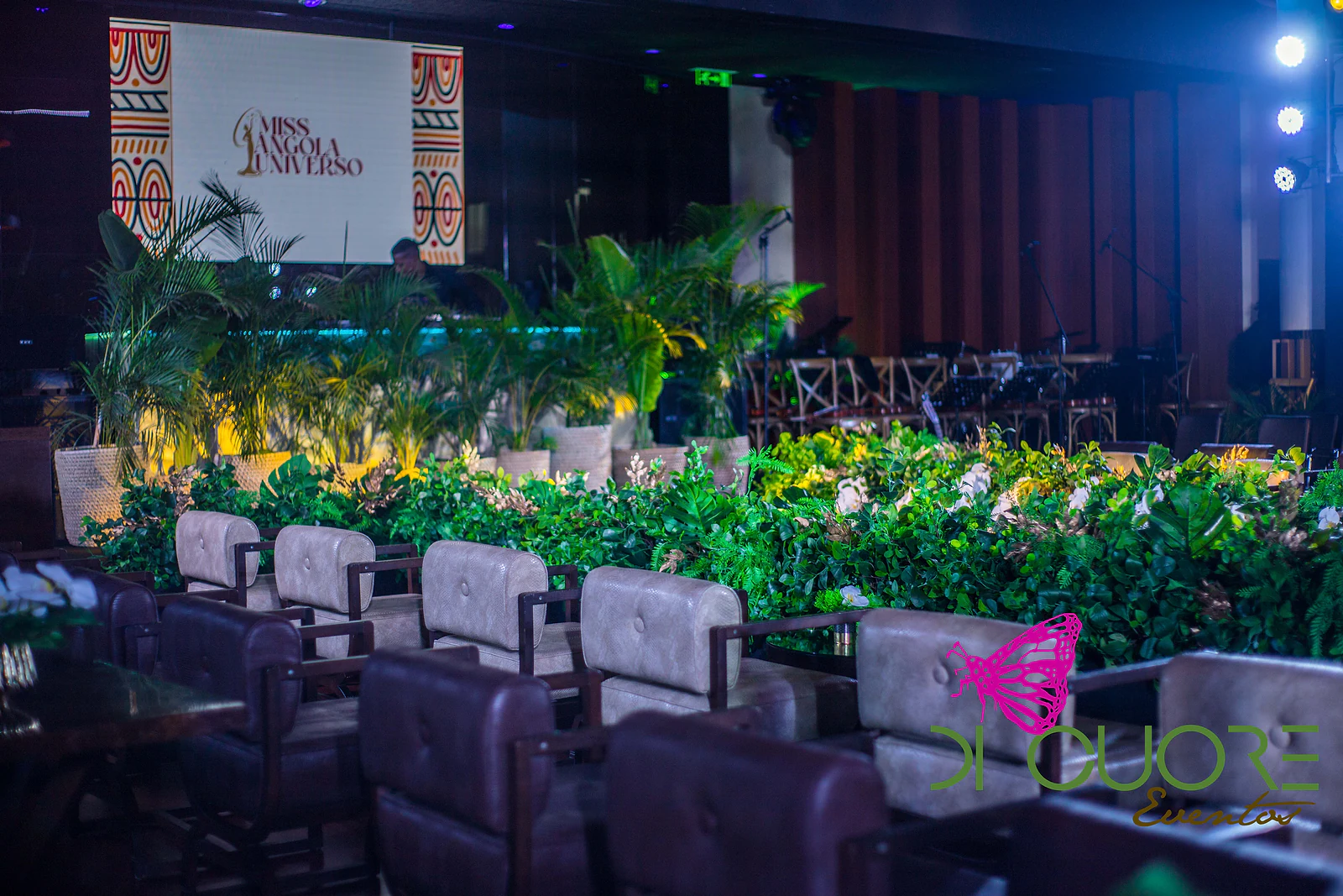 Fila de cadeiras diante de uma sebe de folhagem e flores brancas, com palmeiras e o ecrã do palco ao fundo, sob luz azul