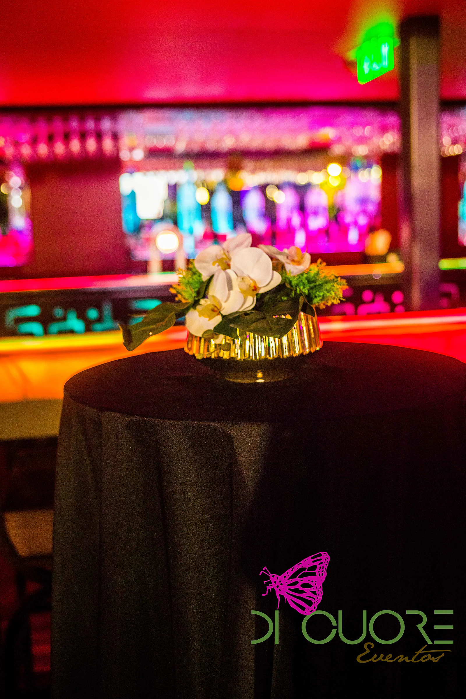 Mesa alta de toalha preta com orquídeas brancas numa taça dourada, diante do bar iluminado em rosa e vermelho