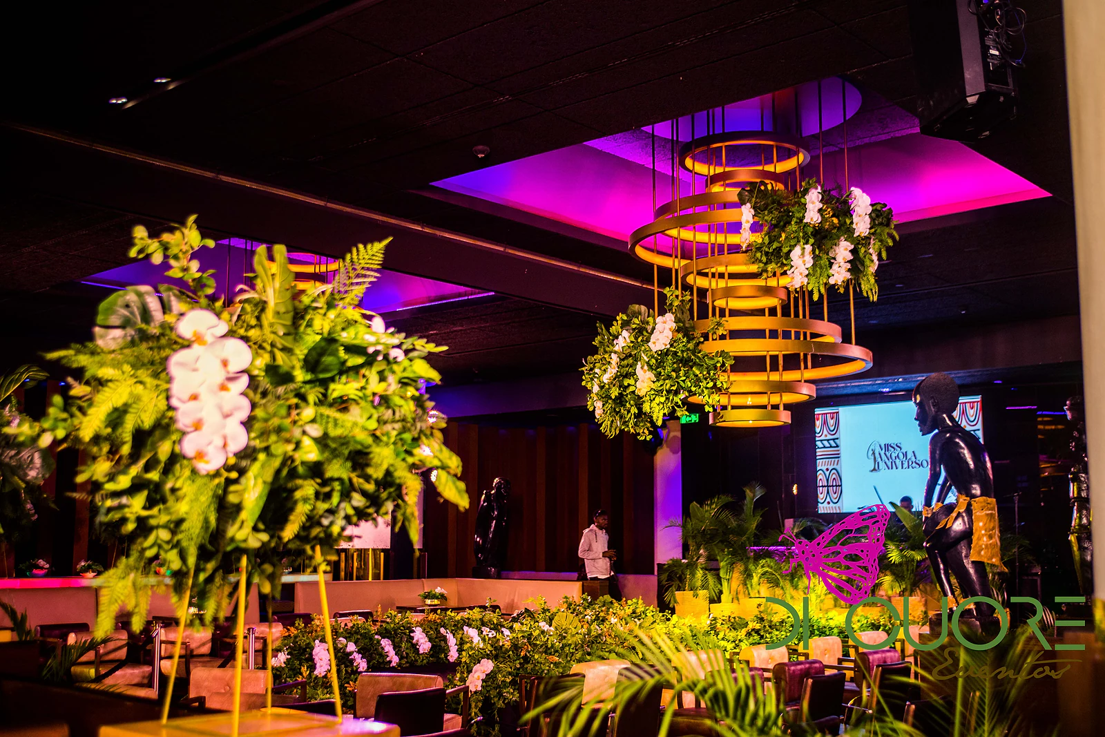 Salão com um arranjo alto de orquídeas brancas e folhagem em primeiro plano e um lustre dourado sob um tecto iluminado a magenta