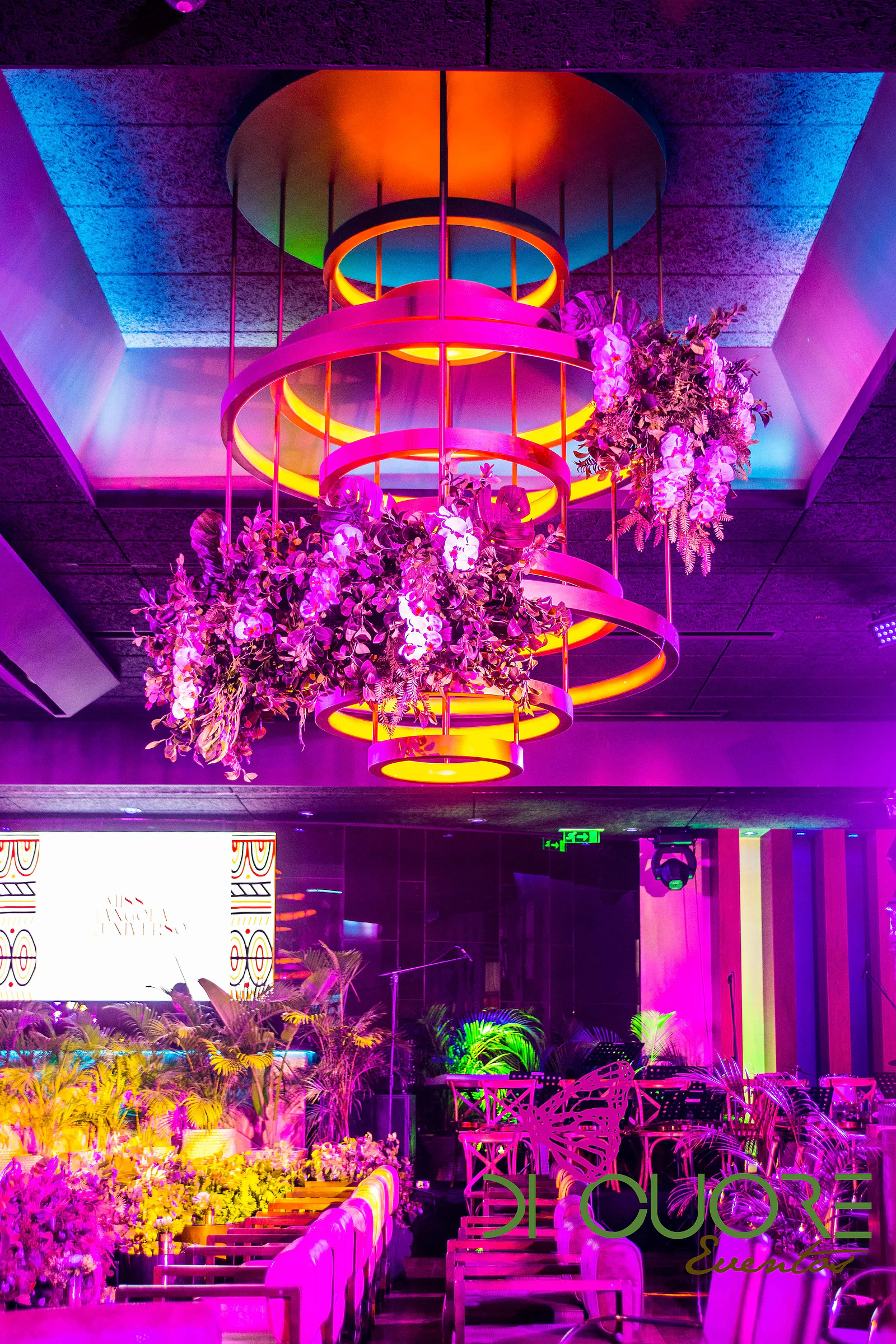 Lustre de aros dourados com flores suspensas sob luz magenta e azul, e as mesas do salão por baixo