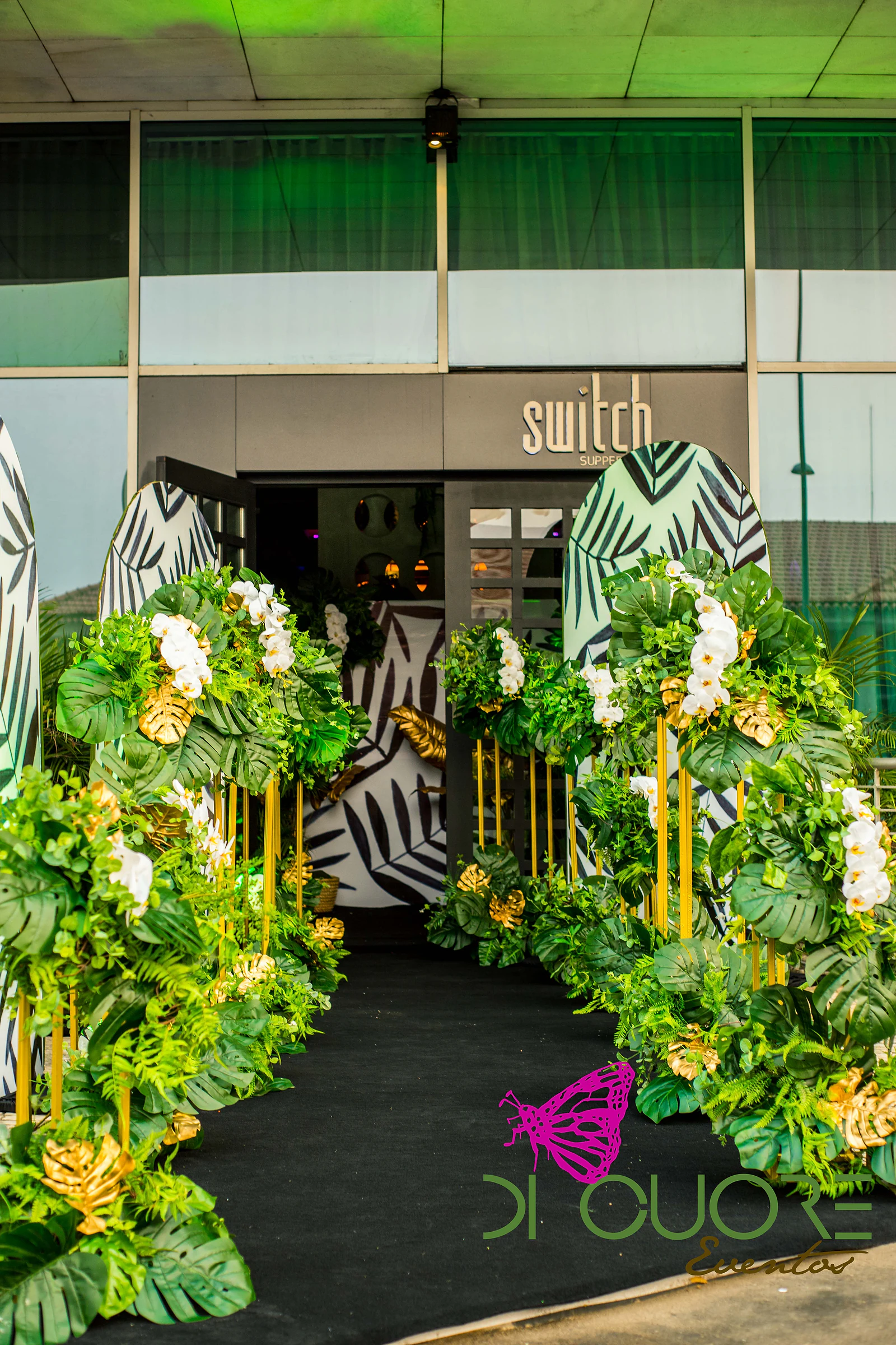 Entrada com passadeira preta entre colunas douradas cobertas de folhas, monsteras e orquídeas brancas, com painéis a preto e branco e um letreiro ao fundo