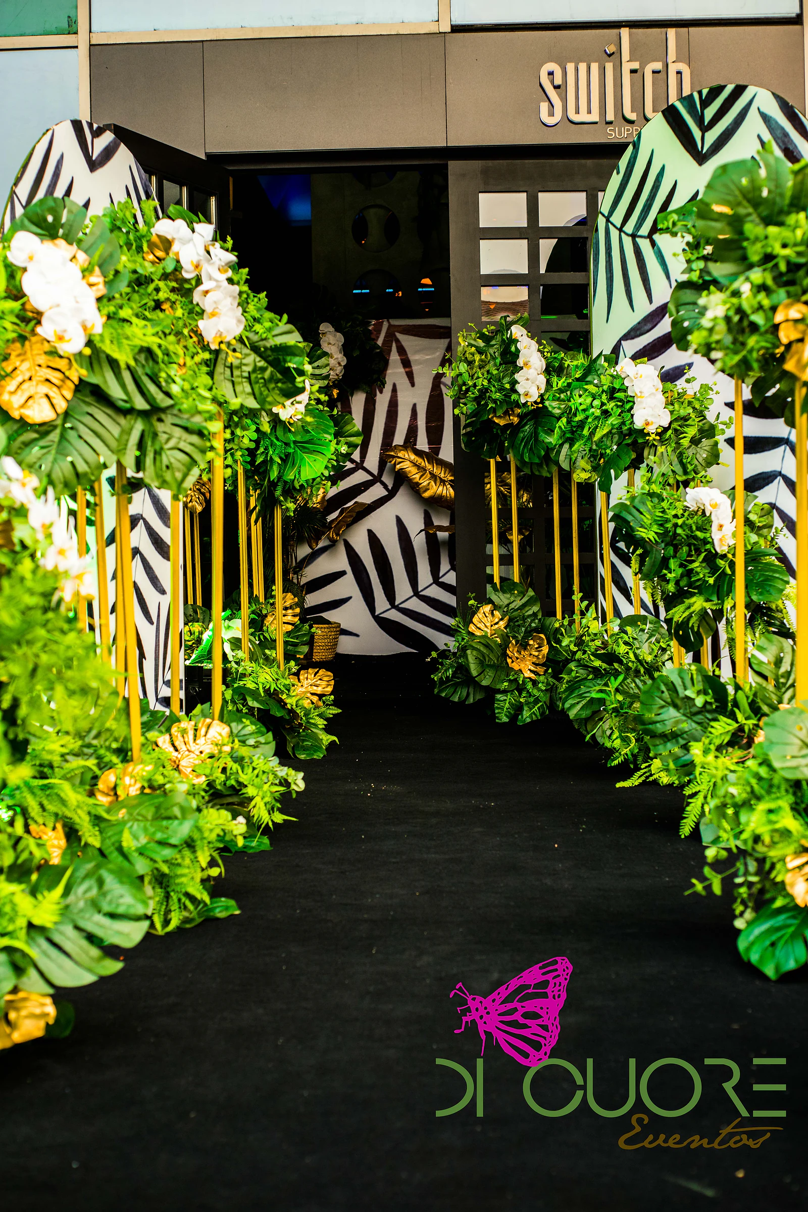 Corredor de passadeira preta entre colunas douradas com monsteras, folhas douradas e orquídeas brancas, diante de painéis estampados a preto e branco