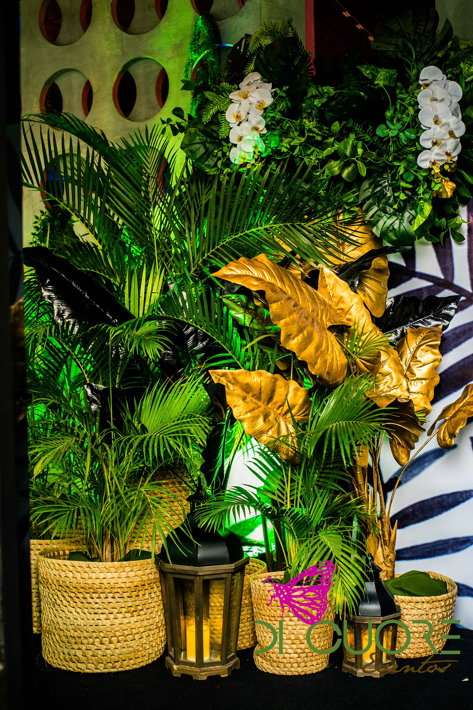 Palmeiras em cestos de palha, folhas douradas, orquídeas brancas e um lampião, diante de um painel de furos redondos iluminado a verde