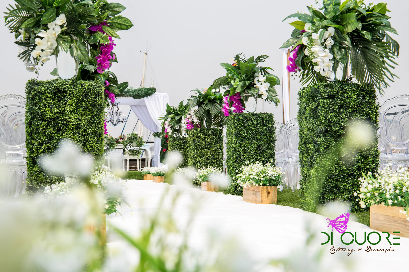 Colunas de buxo com folhagem tropical e orquídeas ao longo da passadeira, com flores brancas desfocadas em primeiro plano e a cabana ao fundo
