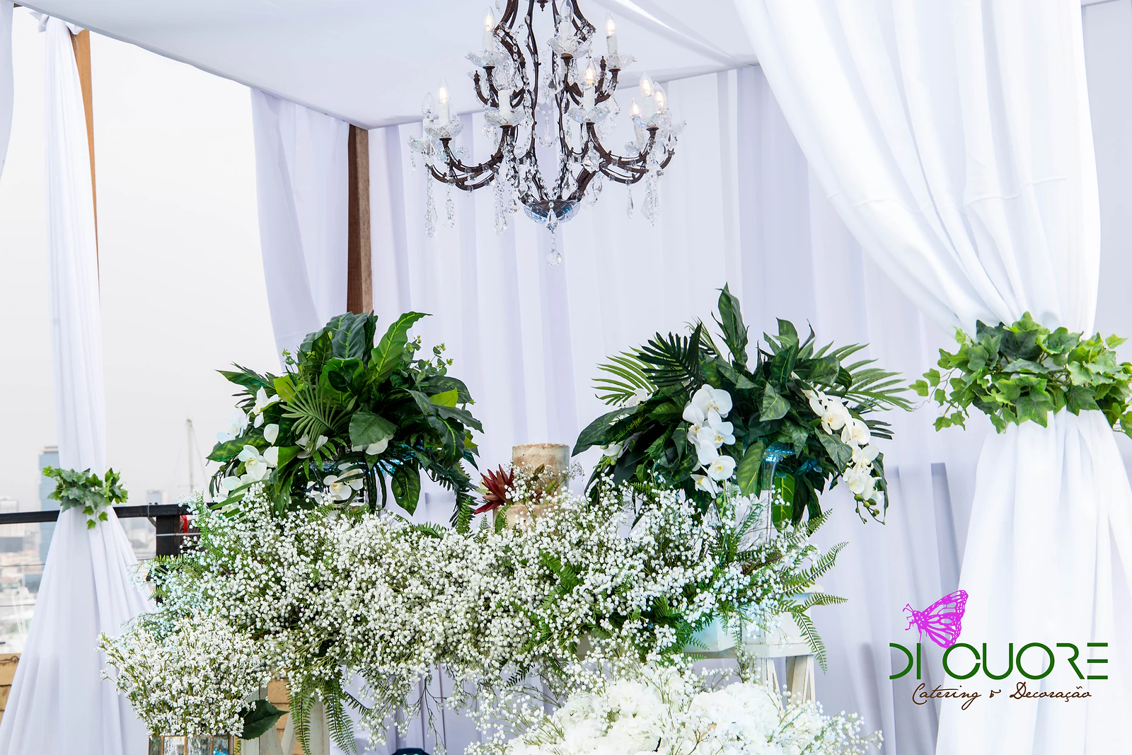 Mesa do bolo coberta de gipsófila sob a cabana de tecido branco e o lustre de cristal, com plantas de folhas grandes e orquídeas brancas