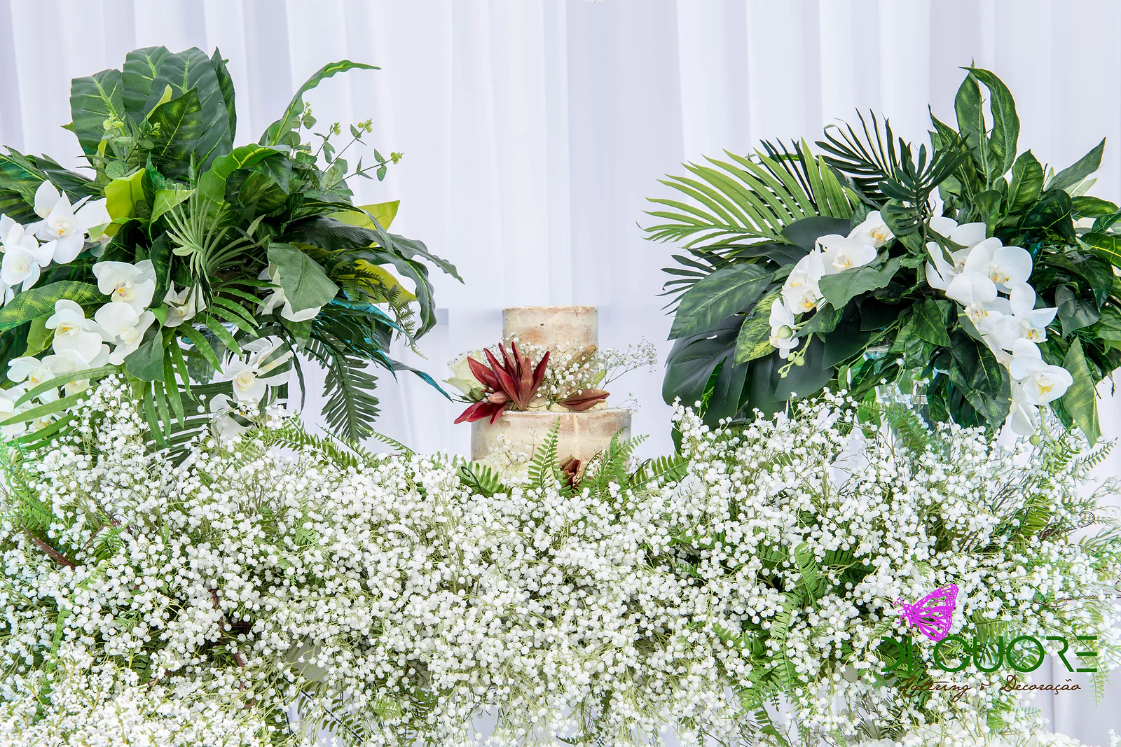 Mesa do bolo envolta numa nuvem de gipsófila, entre arranjos de folhas tropicais e orquídeas brancas diante de cortinados brancos