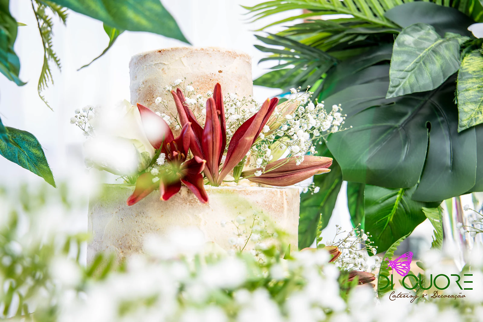 Bolo rústico com flores cor de vinho, entre gipsófila e folhas tropicais grandes