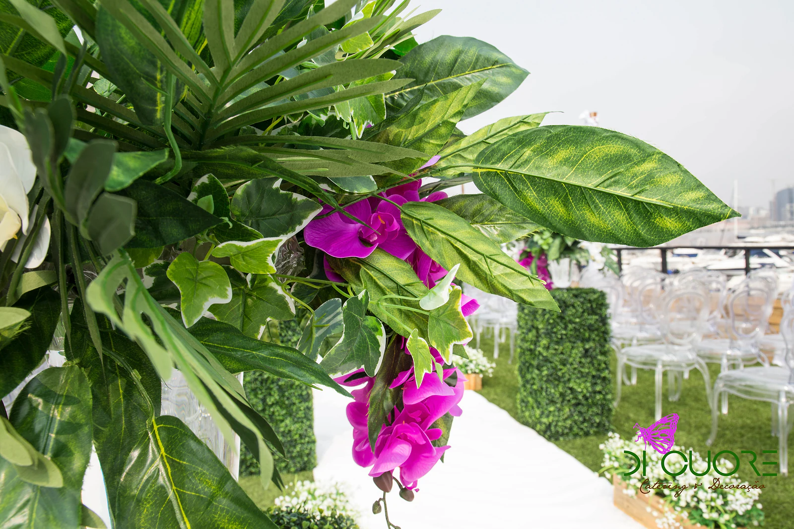 Pormenor de folhas tropicais e orquídeas fúcsia sobre a passadeira, com cadeiras transparentes e uma coluna de buxo ao fundo