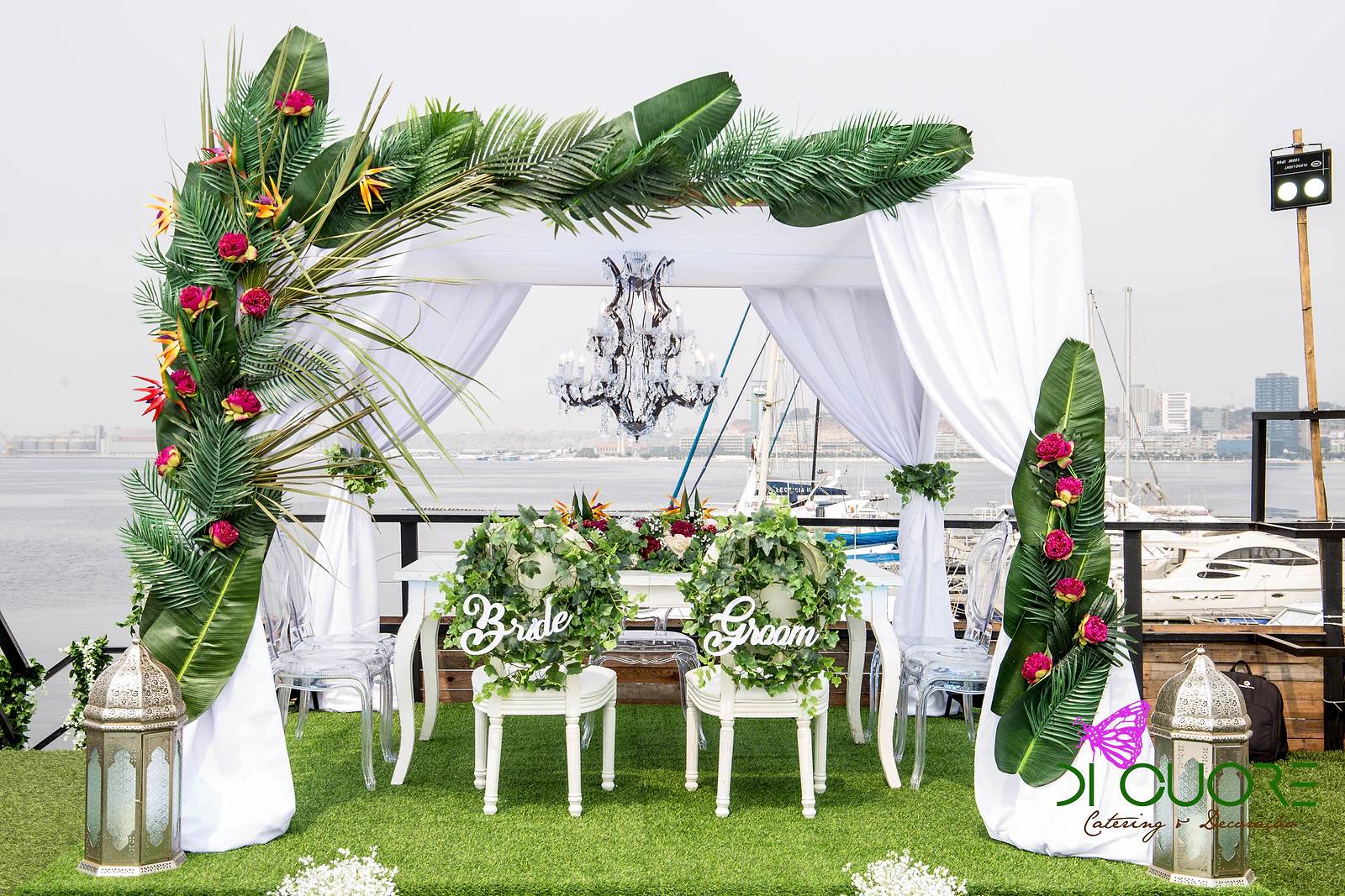 Cabana de tecido branco com lustre de cristal e arco de palmas com flores vermelhas, e as cadeiras brancas dos noivos com grinaldas de hera e os letreiros Bride e Groom