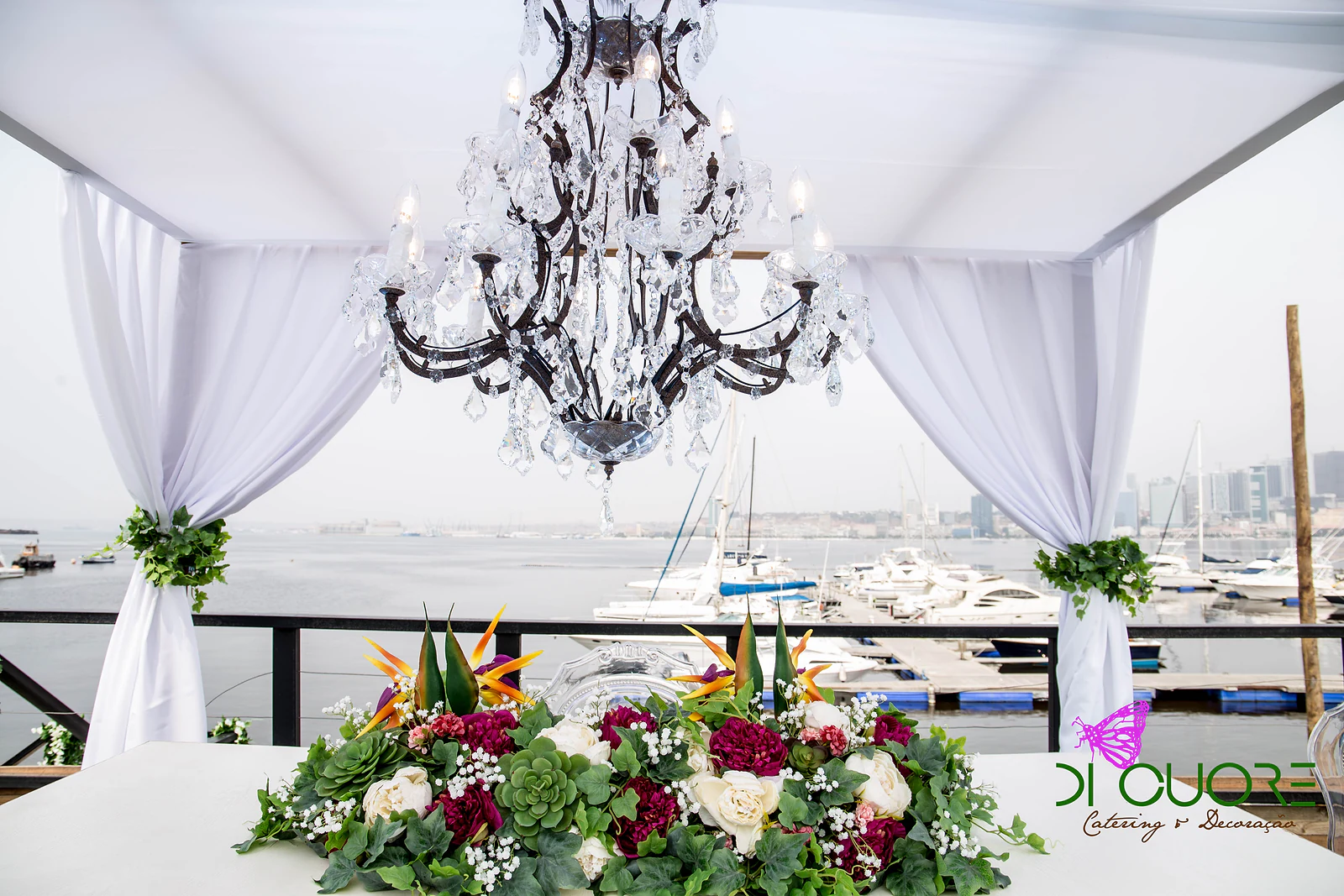 Lustre de cristal sob o tecto de tecido branco, com um arranjo baixo de rosas brancas e cor de vinho, flores tropicais laranja e folhagem sobre a mesa, e os barcos da baía ao fundo