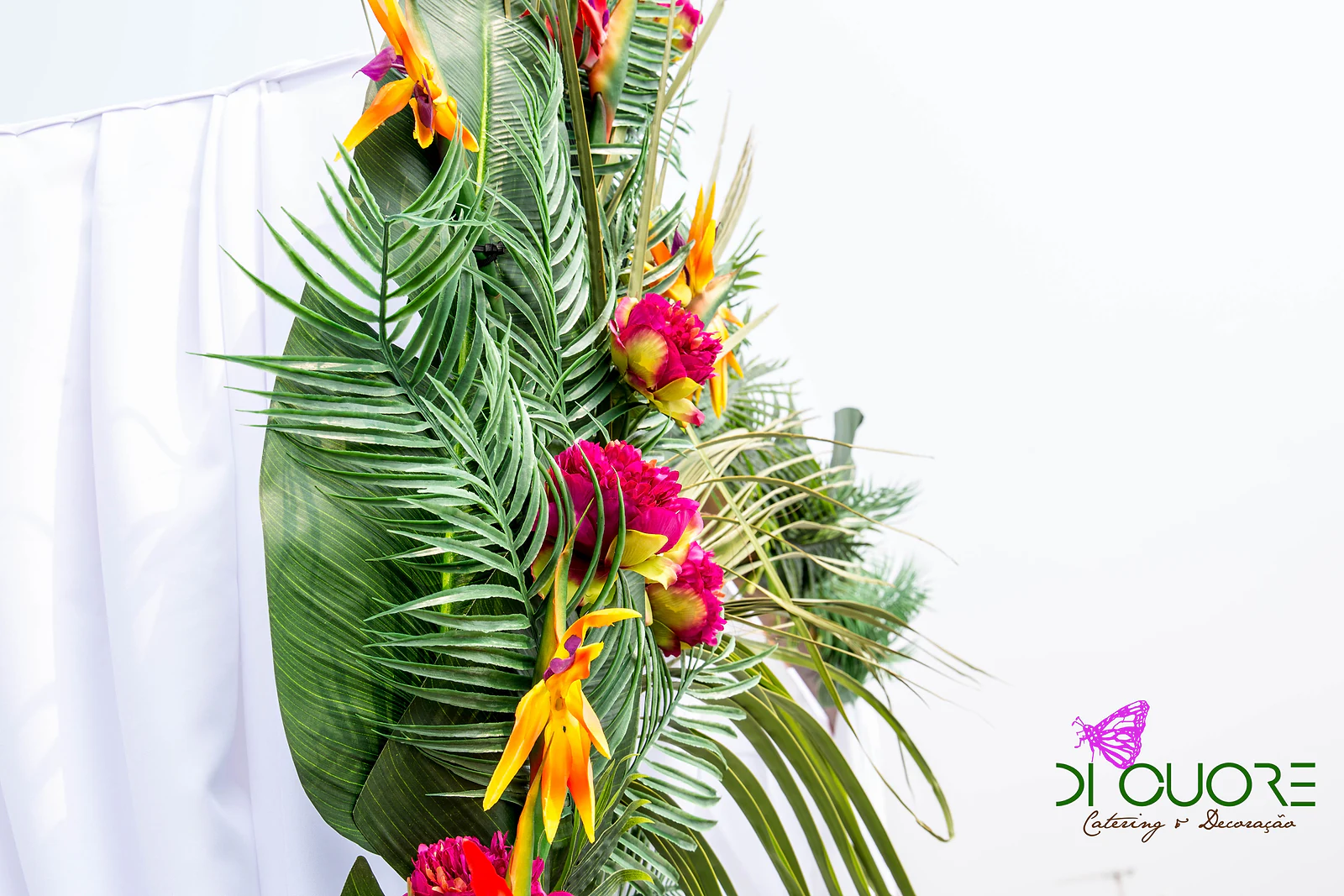 Coluna de folhas de palmeira e de bananeira com flores tropicais laranja e vermelhas, junto a um cortinado branco