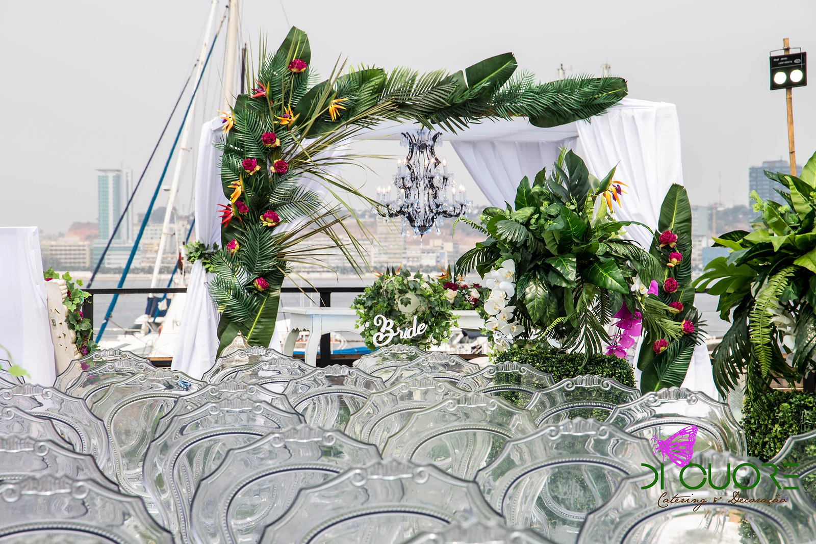 Cadeiras transparentes de espaldar trabalhado em primeiro plano e, ao fundo, o arco de palmas com flores vermelhas junto à cabana branca