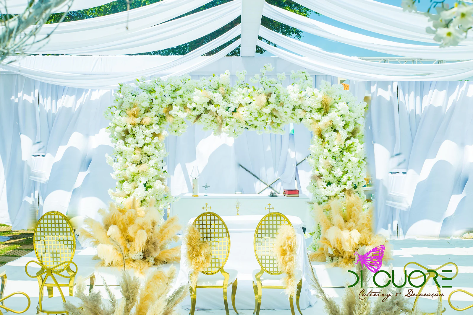 Altar de frente: moldura de flores brancas e pampas sobre a mesa de toalha branca, com duas cadeiras douradas de grelha oval à frente
