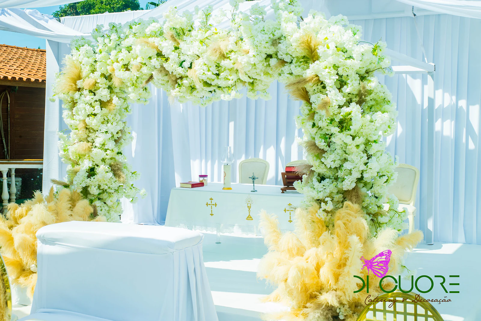 Moldura de flores brancas e plumas douradas sobre o altar, com a mesa de toalha branca e cruzes douradas e cortinados por trás