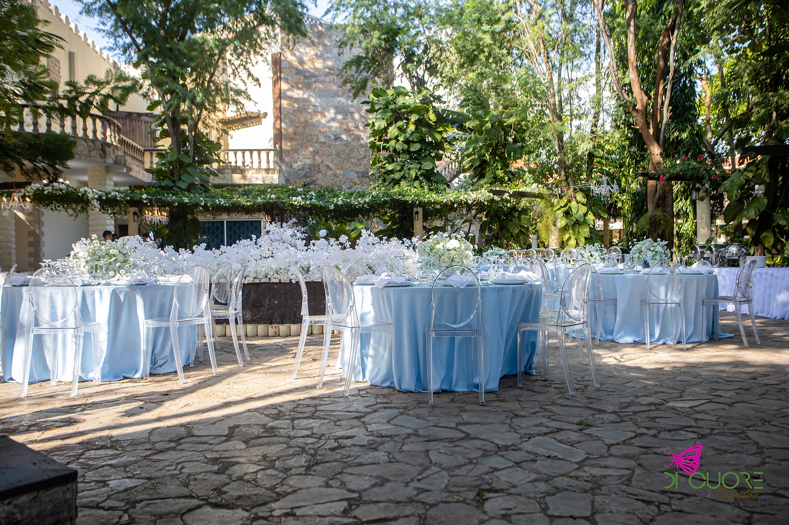 Mesas redondas azul-bebé com cadeiras transparentes diante do canteiro de flores brancas e de uma pérgola de folhagem