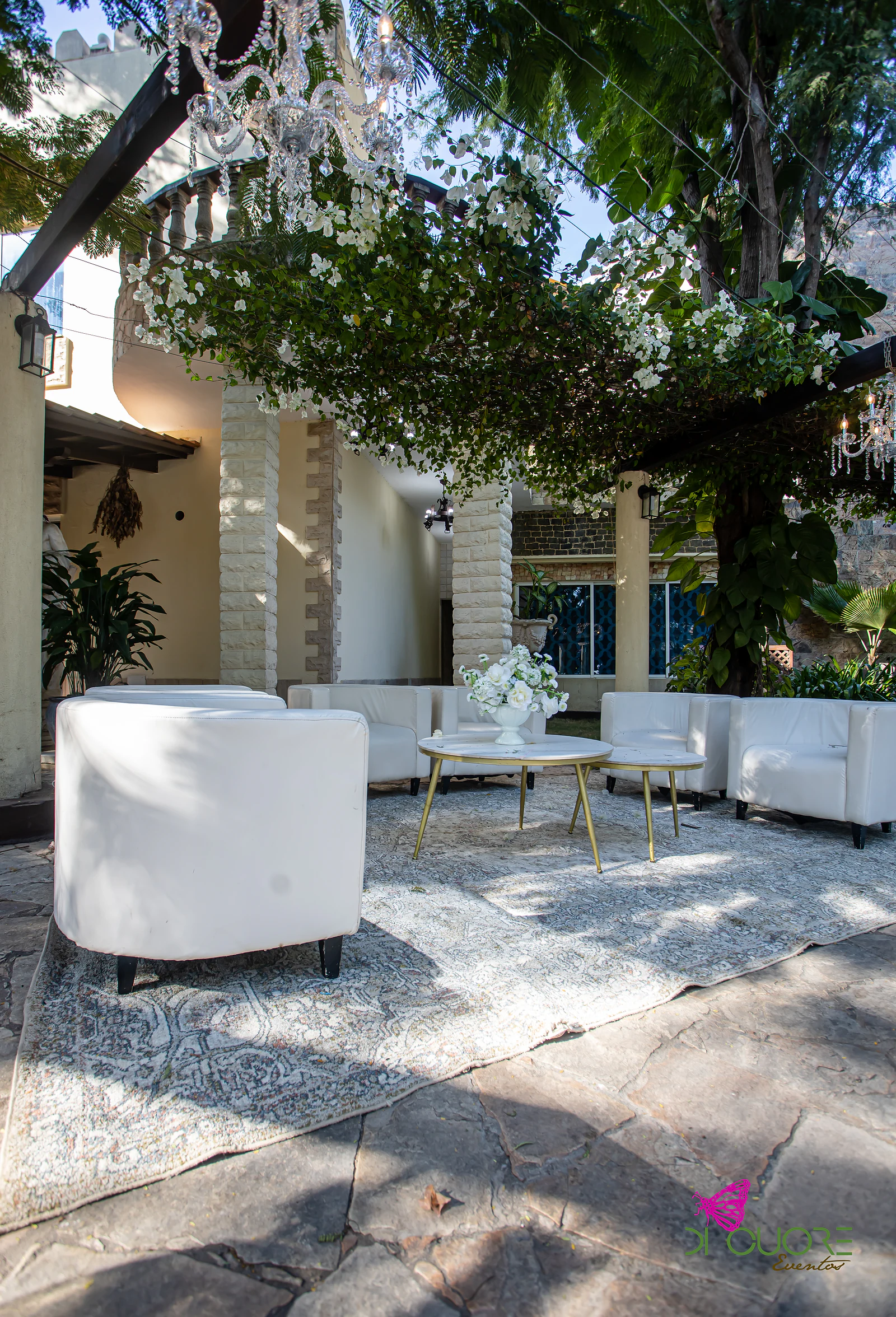 Zona de estar ao ar livre com sofás e cadeiras brancos, mesas douradas, tapete de padrão e uma pérgola coberta de flores brancas