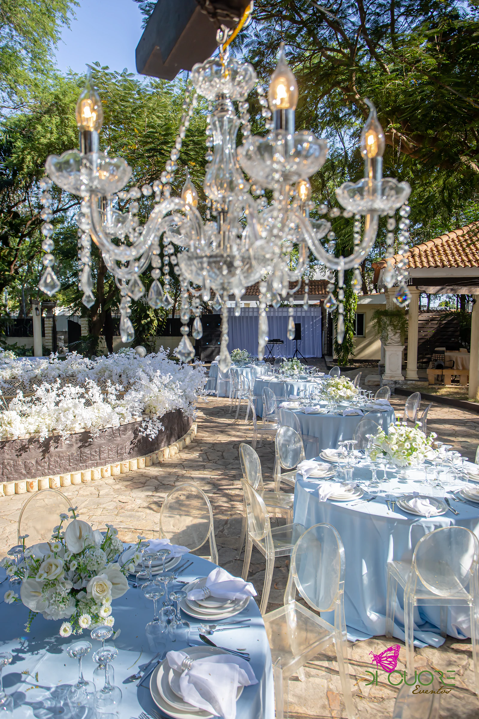 Lustre de cristal com velas sobre mesas redondas de toalha azul-bebé e cadeiras transparentes, junto a um canteiro redondo de flores brancas, com um caramanchão de telhado de barro ao fundo