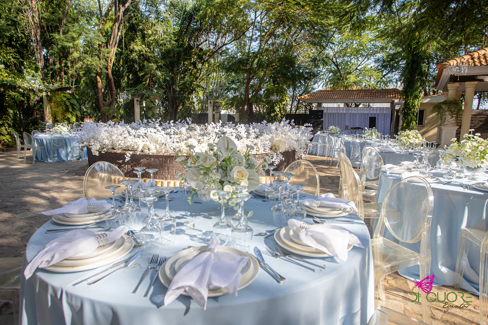 Mesa posta com antúrios e rosas brancas, copos de cristal e guardanapos dobrados sobre toalha azul-bebé, com cortinados cor de areia ao fundo