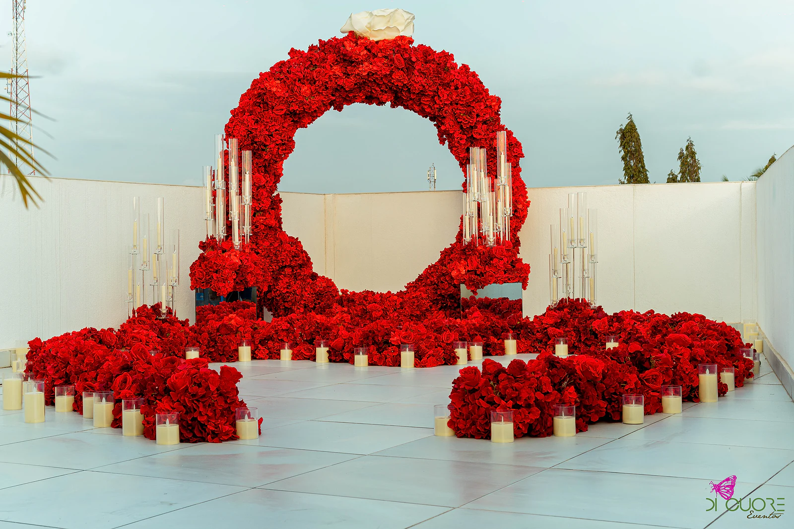 Arco circular de rosas vermelhas com candelabros de vidro e velas, num terraço de paredes brancas, em dia de céu pálido