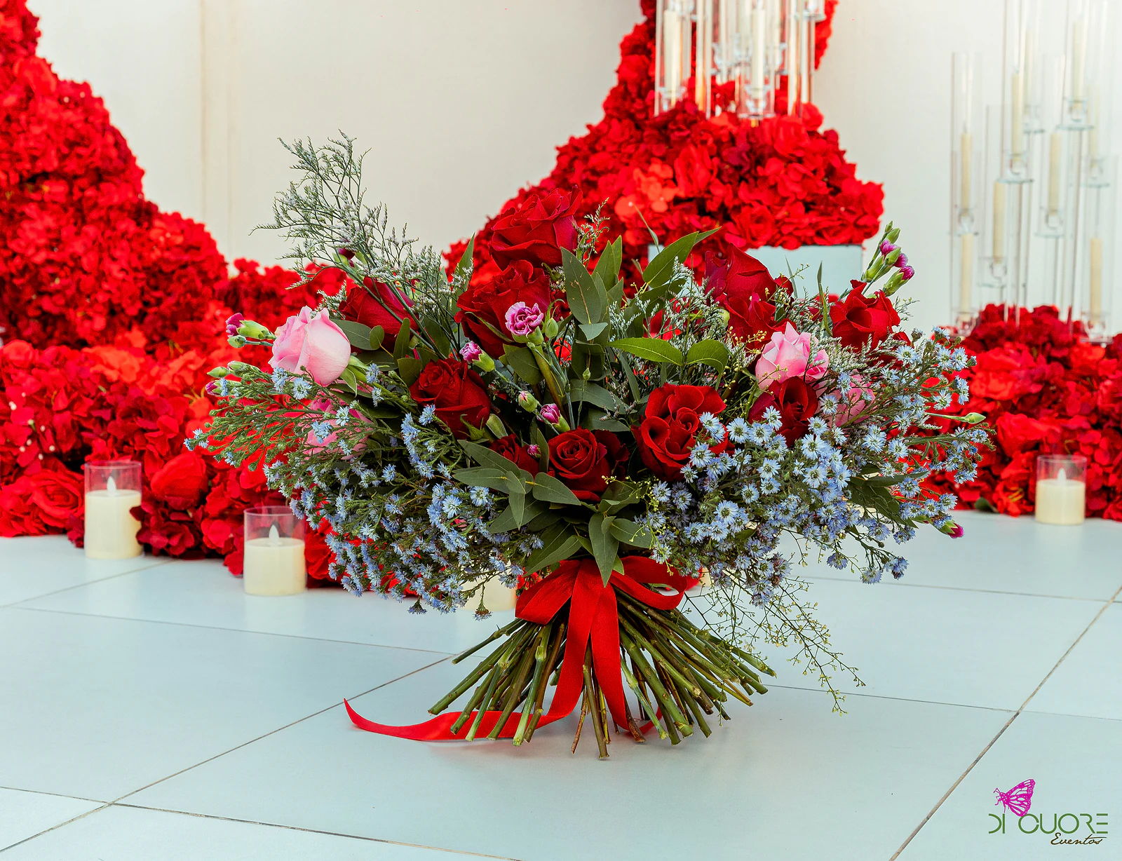 Ramo de rosas e flores azuis com fita vermelha ao nível do piso, em primeiro plano, com candelabros de vidro e maciços de rosas ao fundo