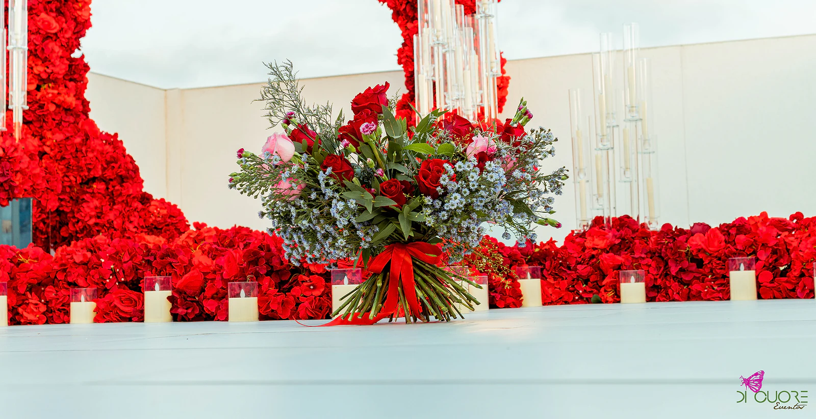 Ramo de rosas e flores azuis sobre o piso claro, com velas em copos de vidro e o arco de rosas a surgir ao fundo