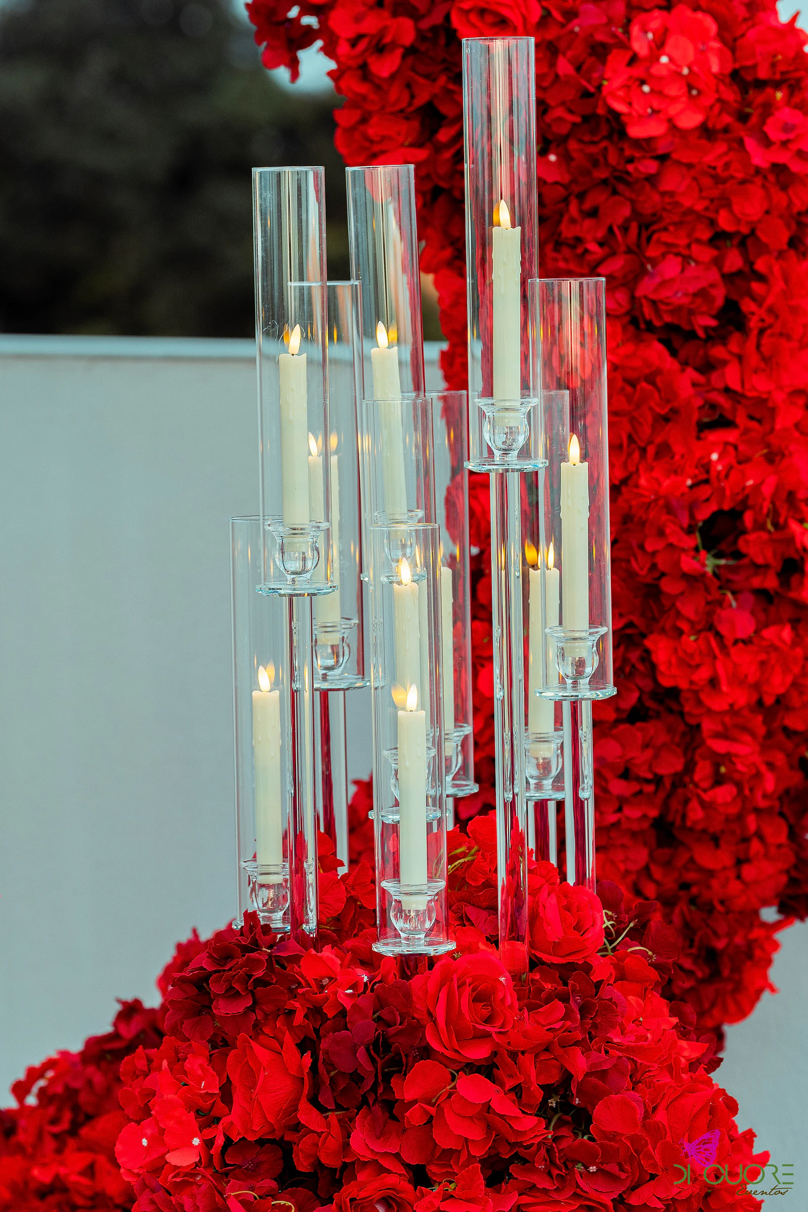 Candelabros de vidro com velas altas acesas sobre um maciço de rosas vermelhas, junto ao arco de flores
