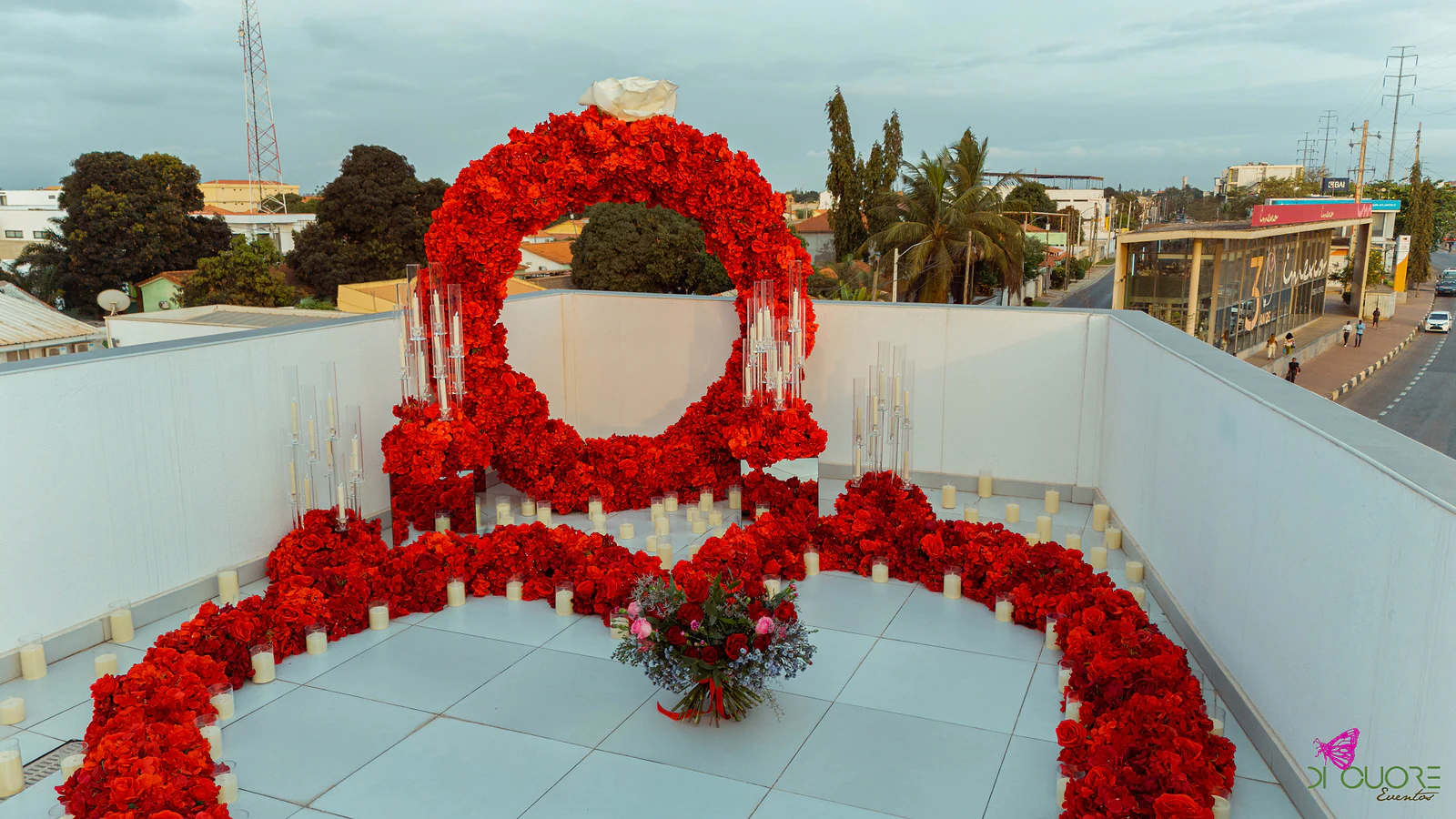Vista elevada do terraço de dia, com o arco de rosas vermelhas rematado por uma rosa branca, a fita de rosas e o ramo ao centro, sobre a cidade