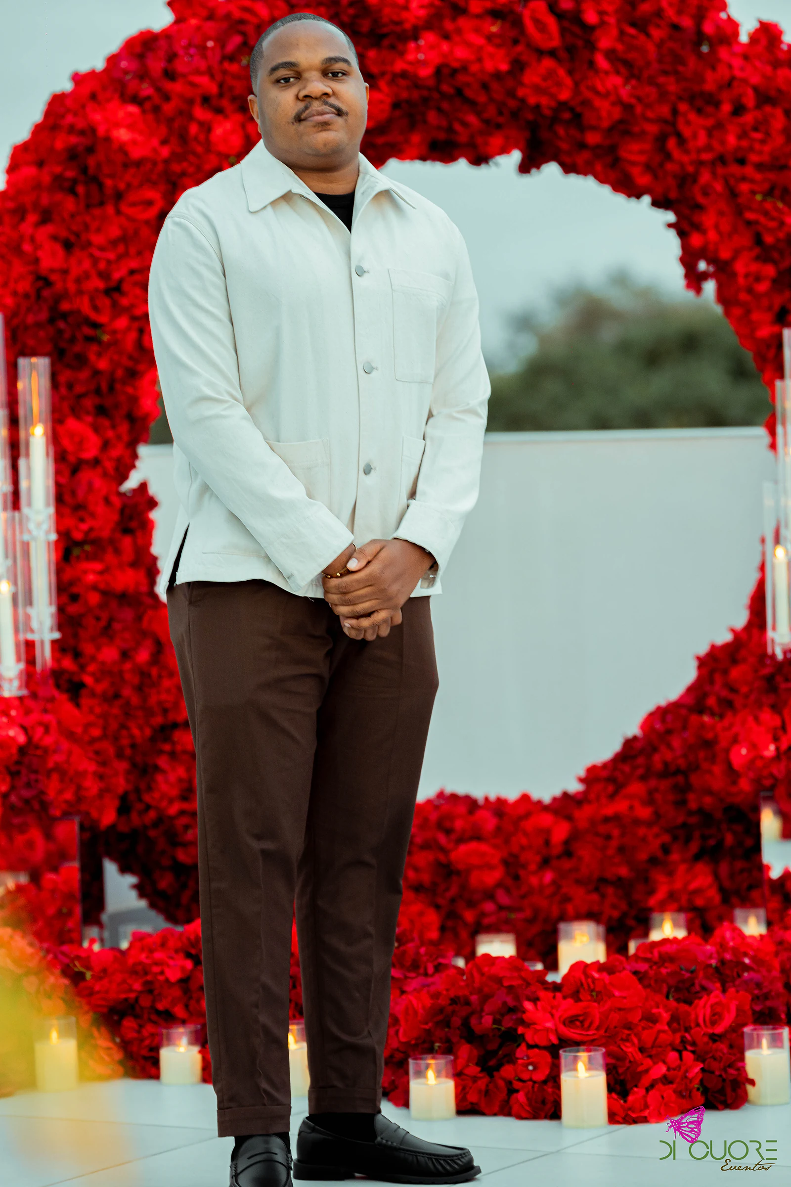 Homem de casaco claro e calças castanhas, de pé diante do arco de rosas vermelhas, com velas ao fundo