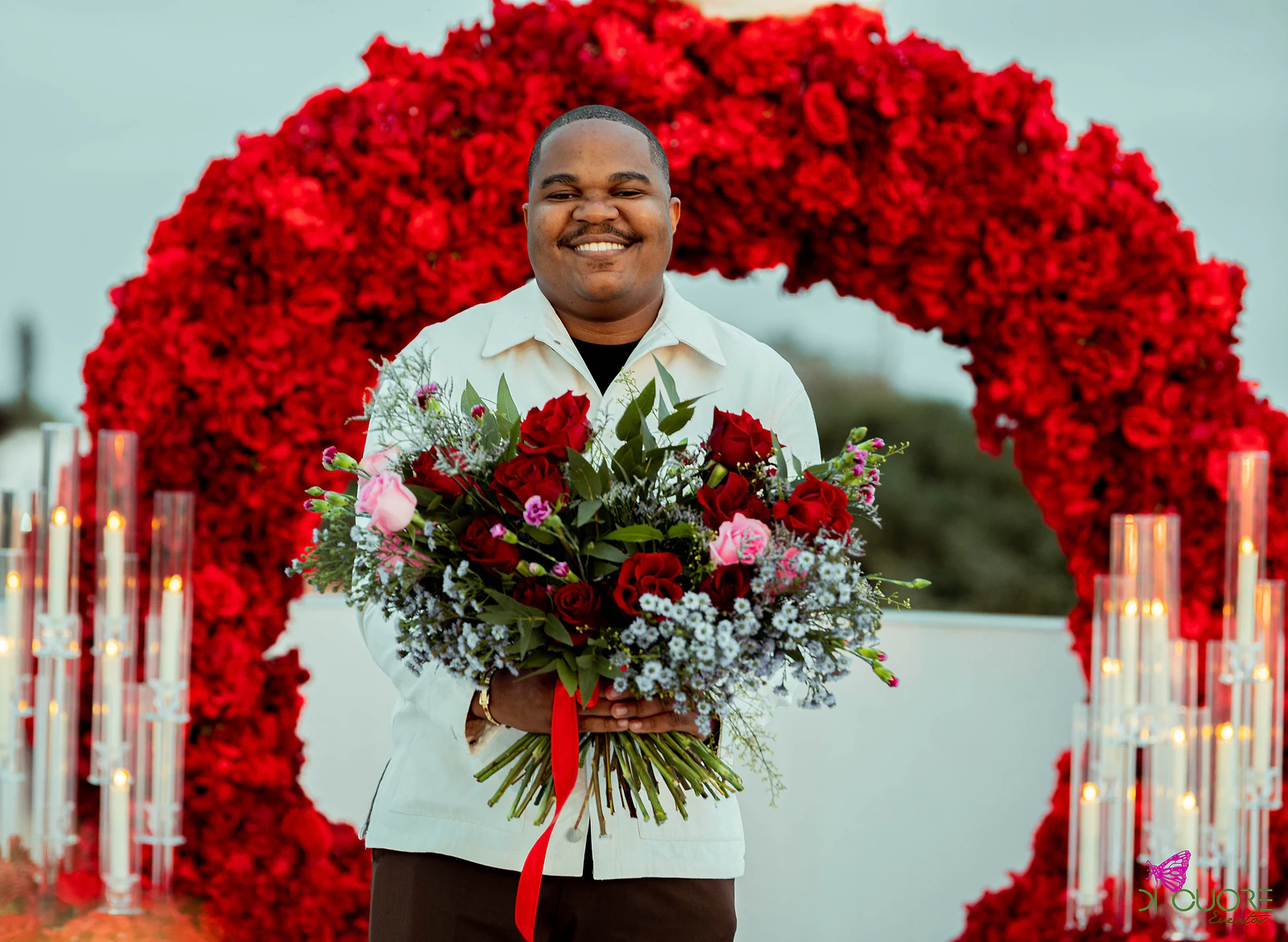 Homem de casaco claro a sorrir, com um ramo de rosas vermelhas e cor-de-rosa e flores azuis, diante do arco de rosas