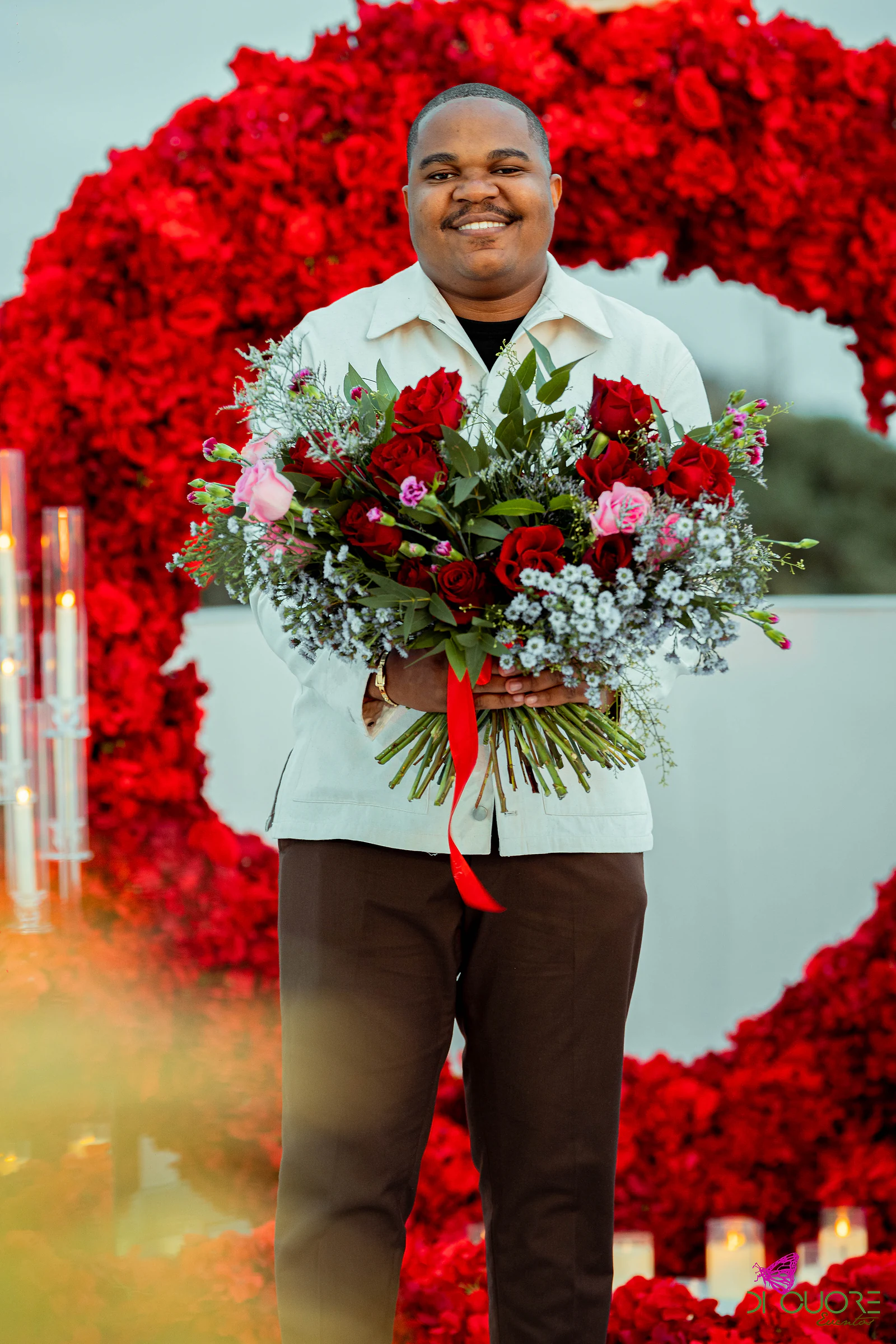 Homem de casaco claro a sorrir, com um grande ramo de rosas e flores azuis, diante do arco de rosas ao entardecer