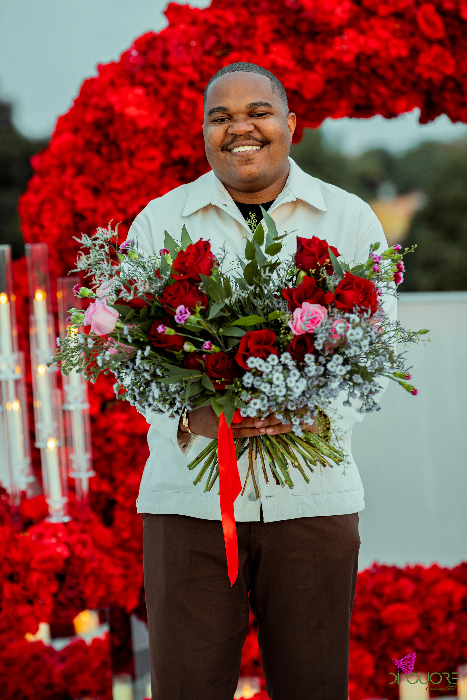 Homem de casaco claro a sorrir, com um grande ramo de rosas e flores azuis, diante do arco de rosas e de candelabros