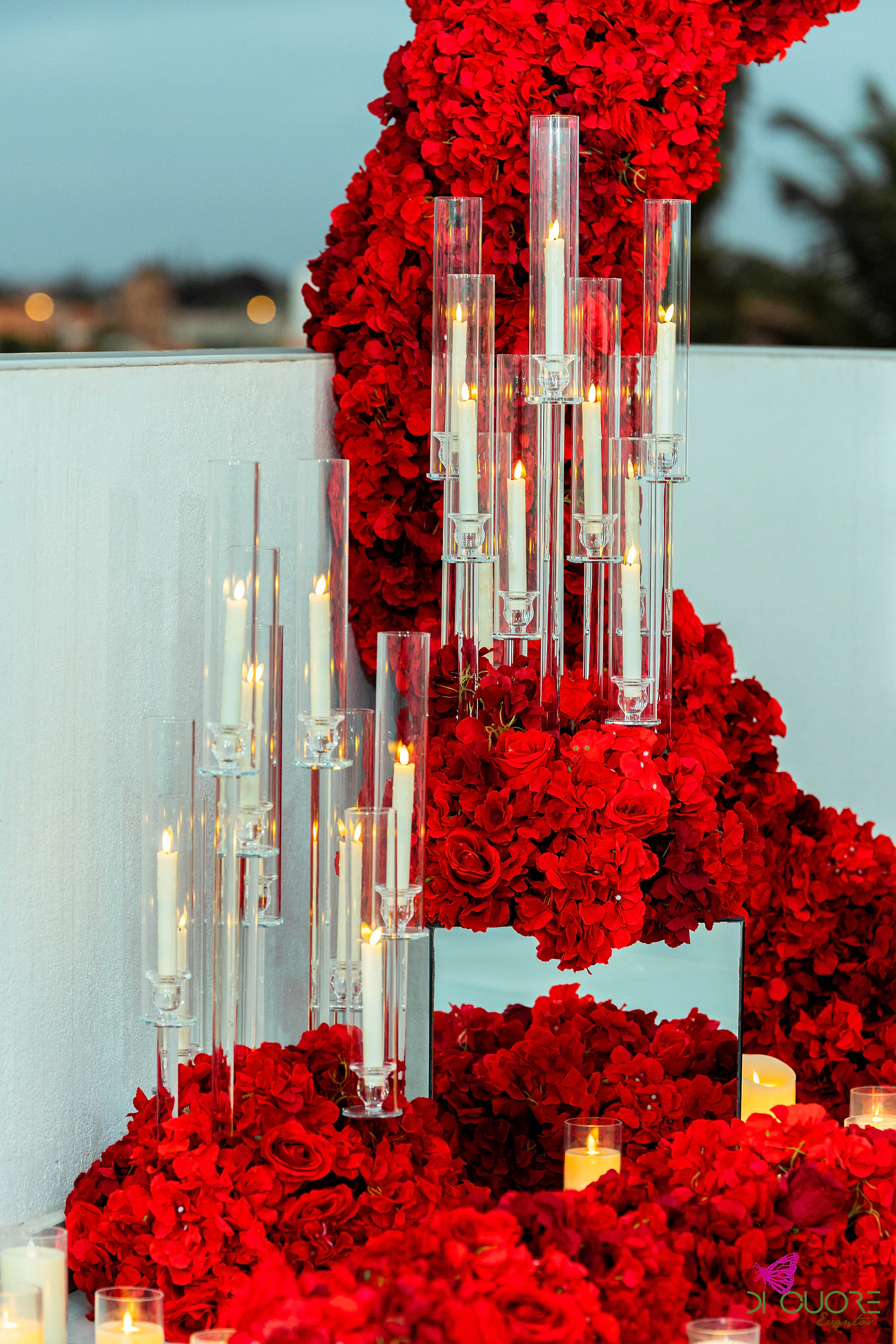 Candelabros de vidro com velas altas junto a uma coluna ondulada de flores vermelhas, com um espelho entre as rosas, ao entardecer